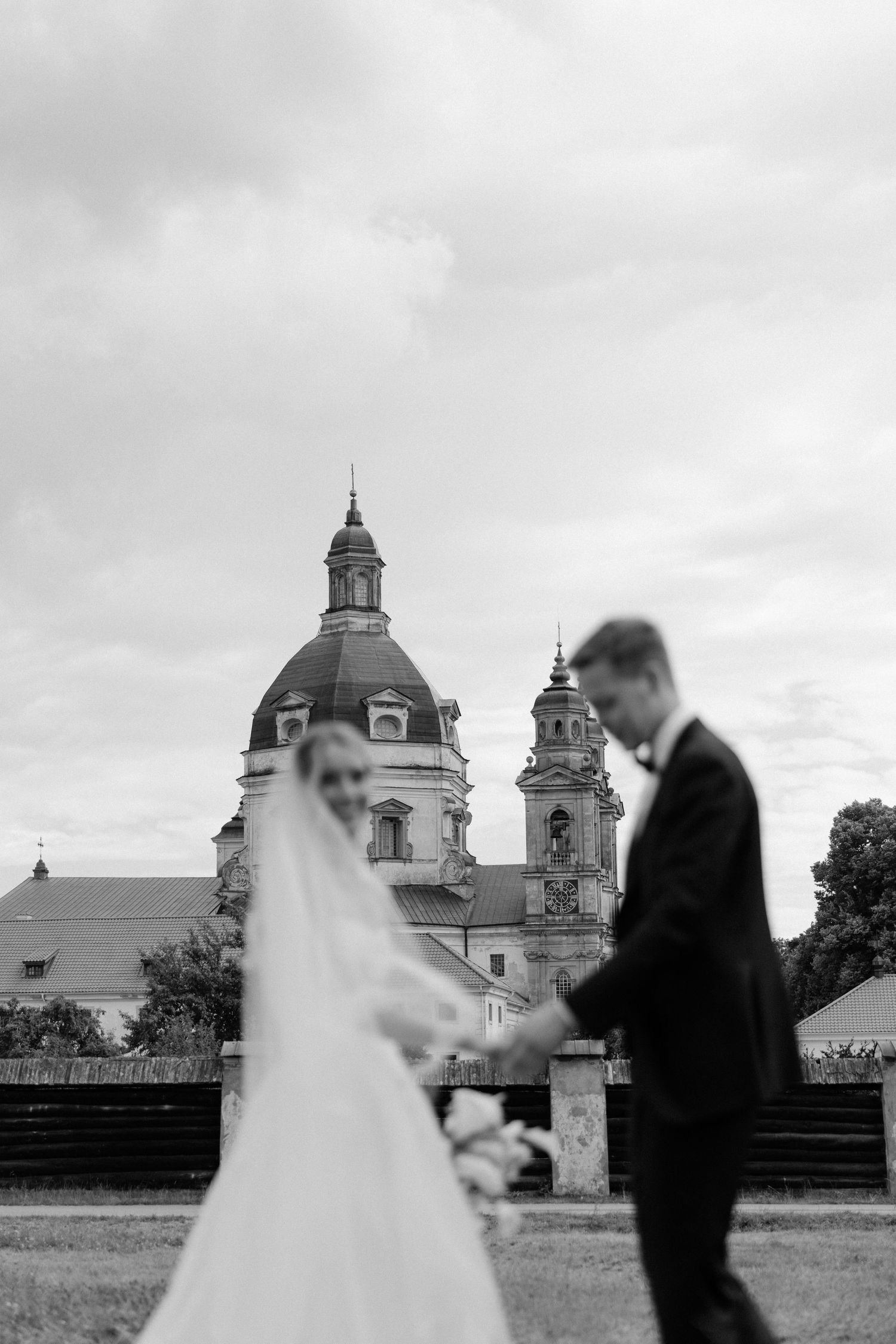 Black and white photo of bride and groom holding hands with a grand domed church building in background.