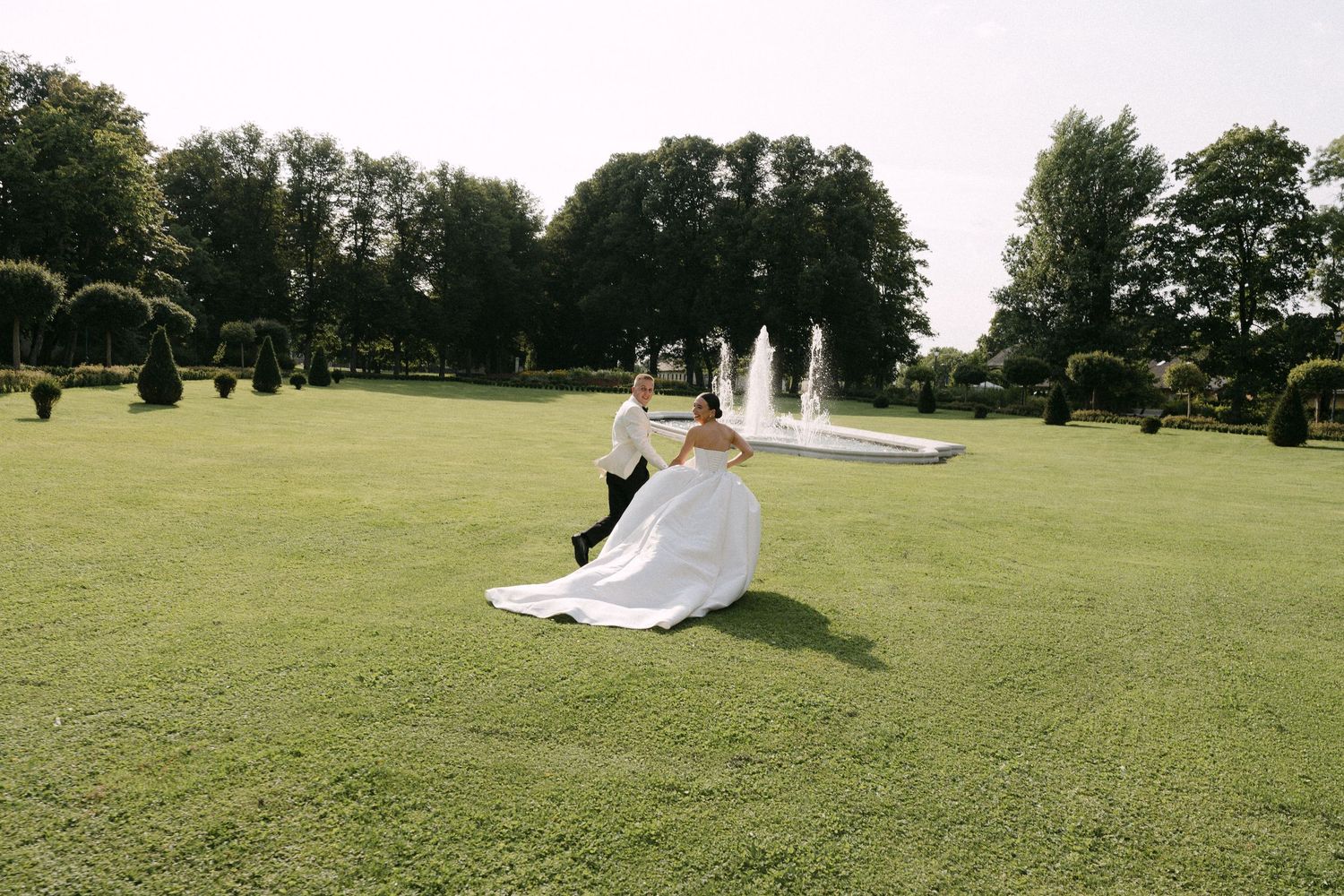 Groom spinning bride in flowing white wedding dress on a manicured green lawn with fountains behind them.