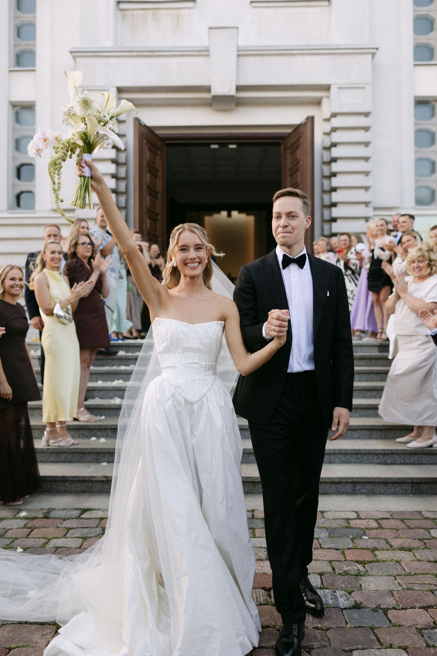 Joyful newlyweds exit church steps, bride in strapless white gown raising bouquet, groom in black tuxedo, guests celebrating.