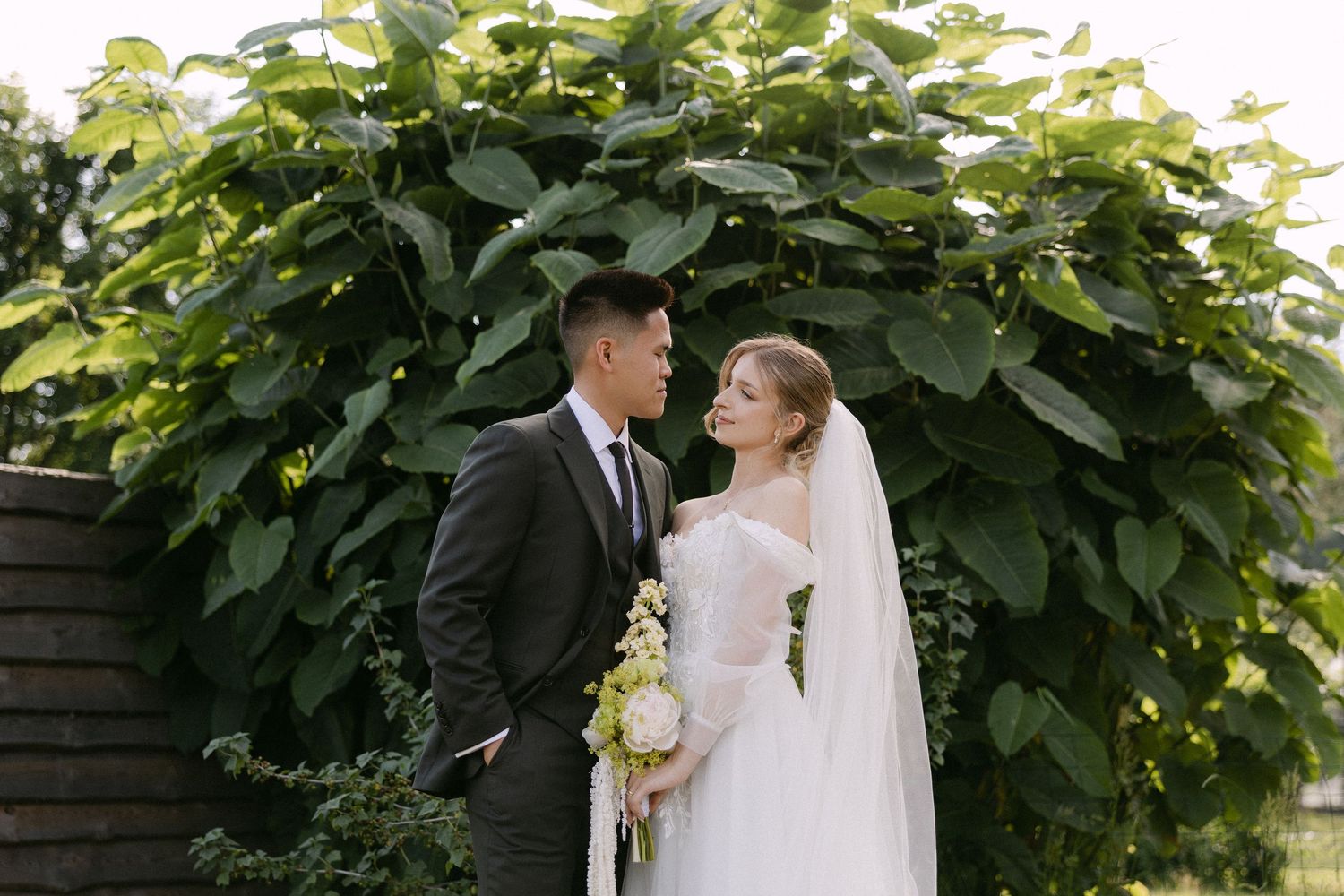 Bride and groom share a romantic gaze beneath a lush green vine archway outdoors.