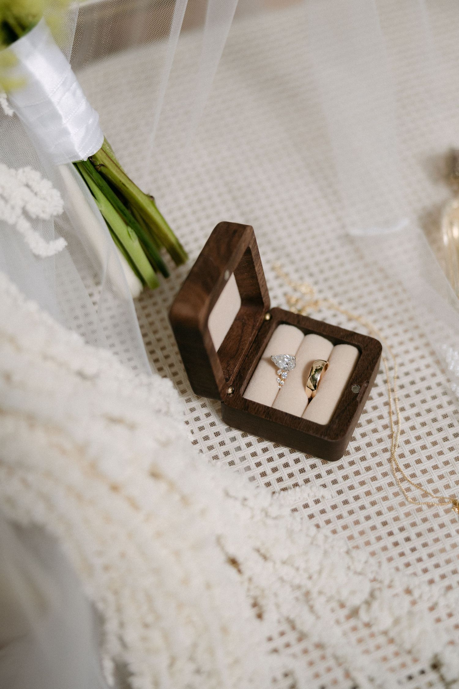 Wedding rings in a wooden box with diamond engagement ring and gold band beside a bridal bouquet and veil.