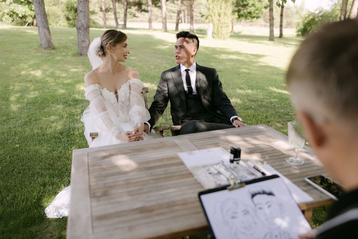 Smiling bride and groom signing marriage documents at a wooden table during outdoor wedding ceremony.