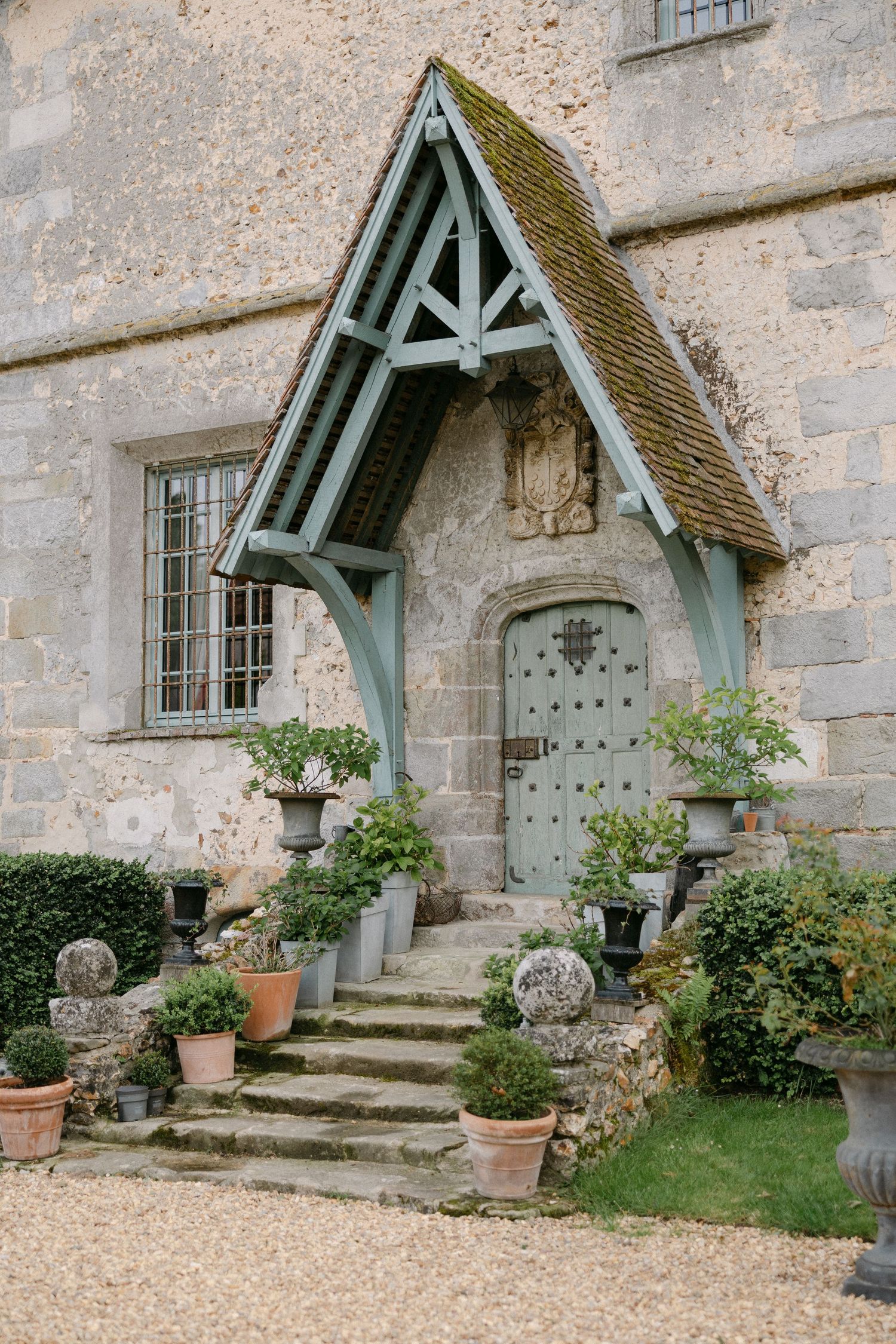 Charming stone cottage entrance with a wooden porch, moss-covered roof, and potted plants on steps.
