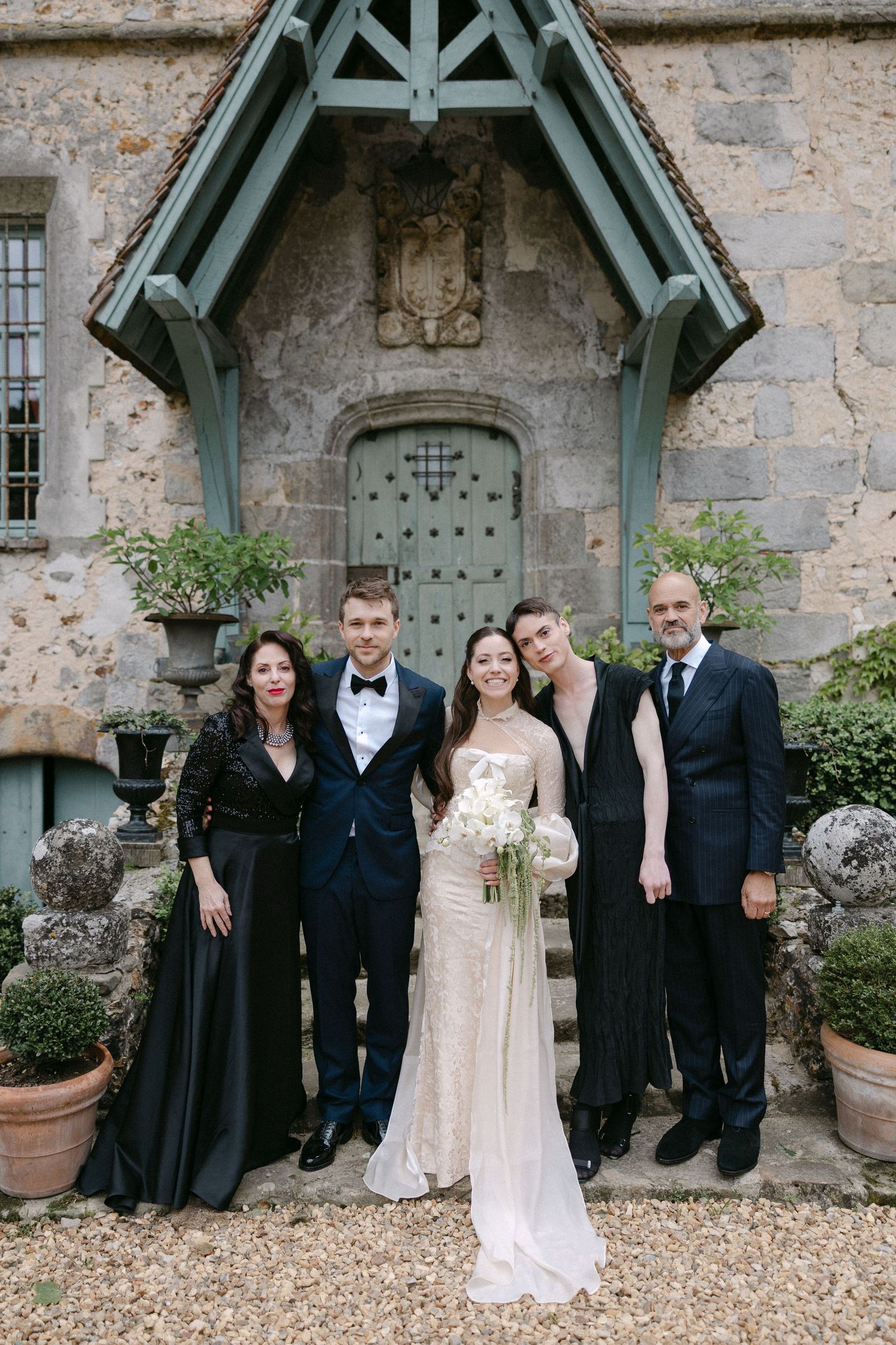 Wedding party of five poses before a stone chateau entrance, bride holding white bouquet in sequin gown.