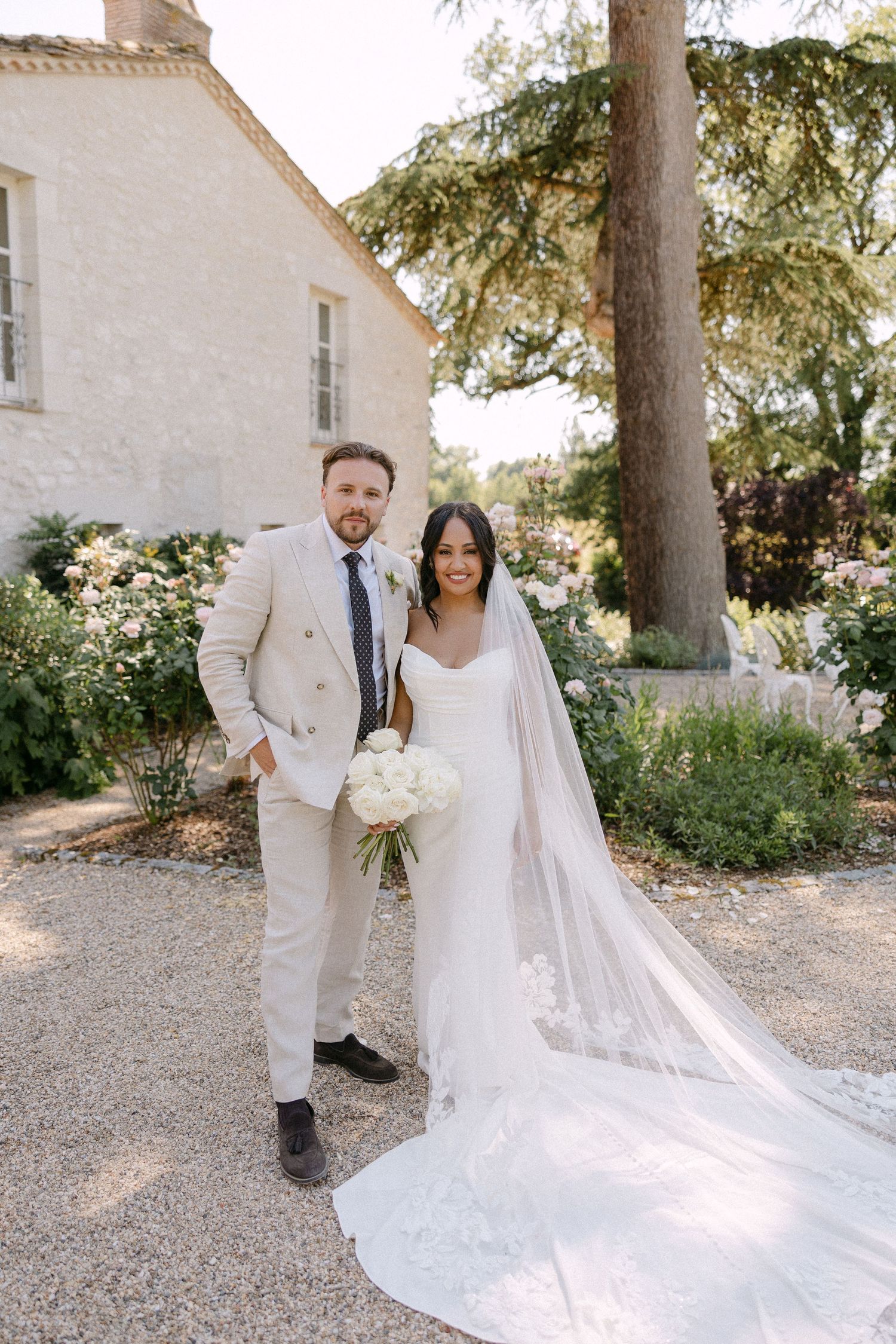 Bride in white gown with lace veil and groom in beige suit posing in a French chateau garden.