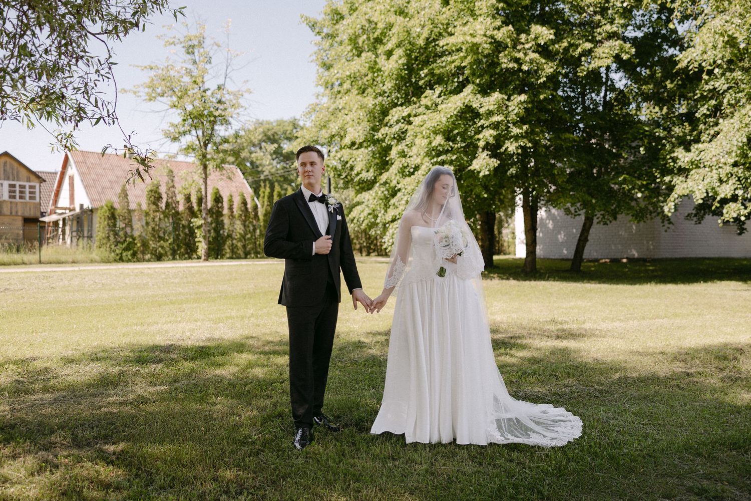 Bride in white gown and groom in black tuxedo hold hands in sunny green garden on wedding day.