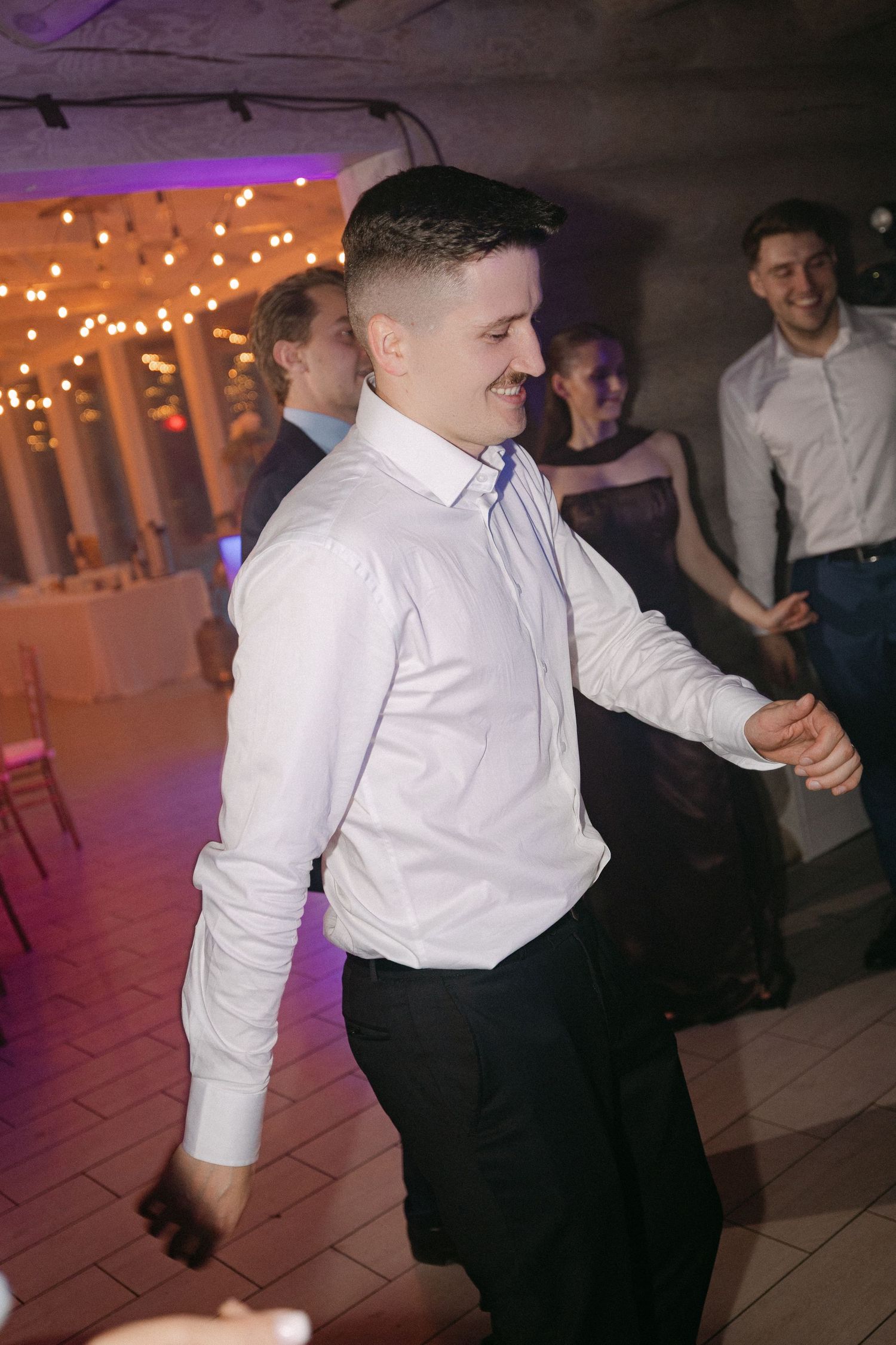 Groom dancing joyfully in white shirt at wedding reception with string lights and guests in background.