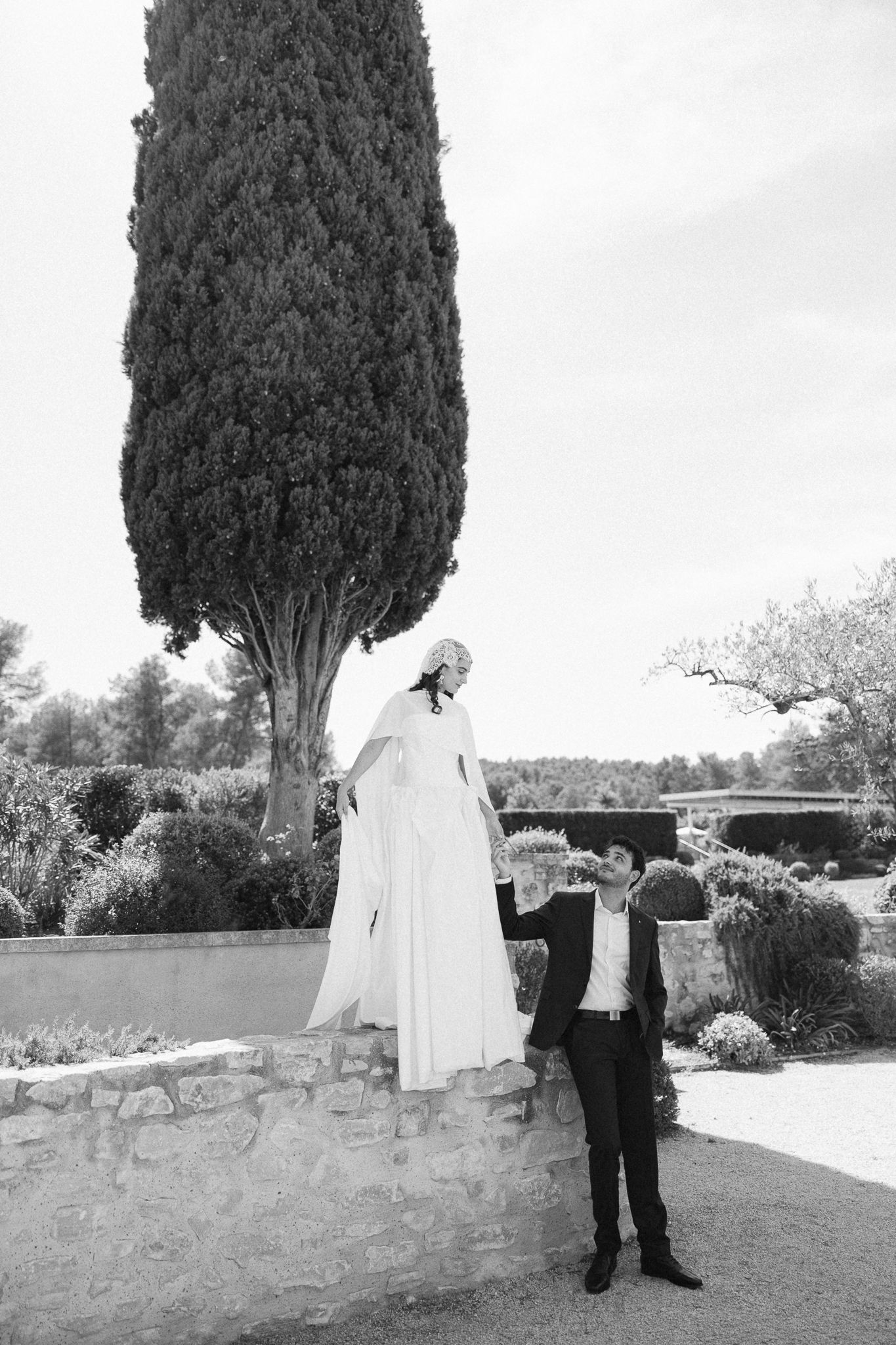Black and white photo of bride in white gown standing on stone wall with groom in suit beside tall cypress tree.