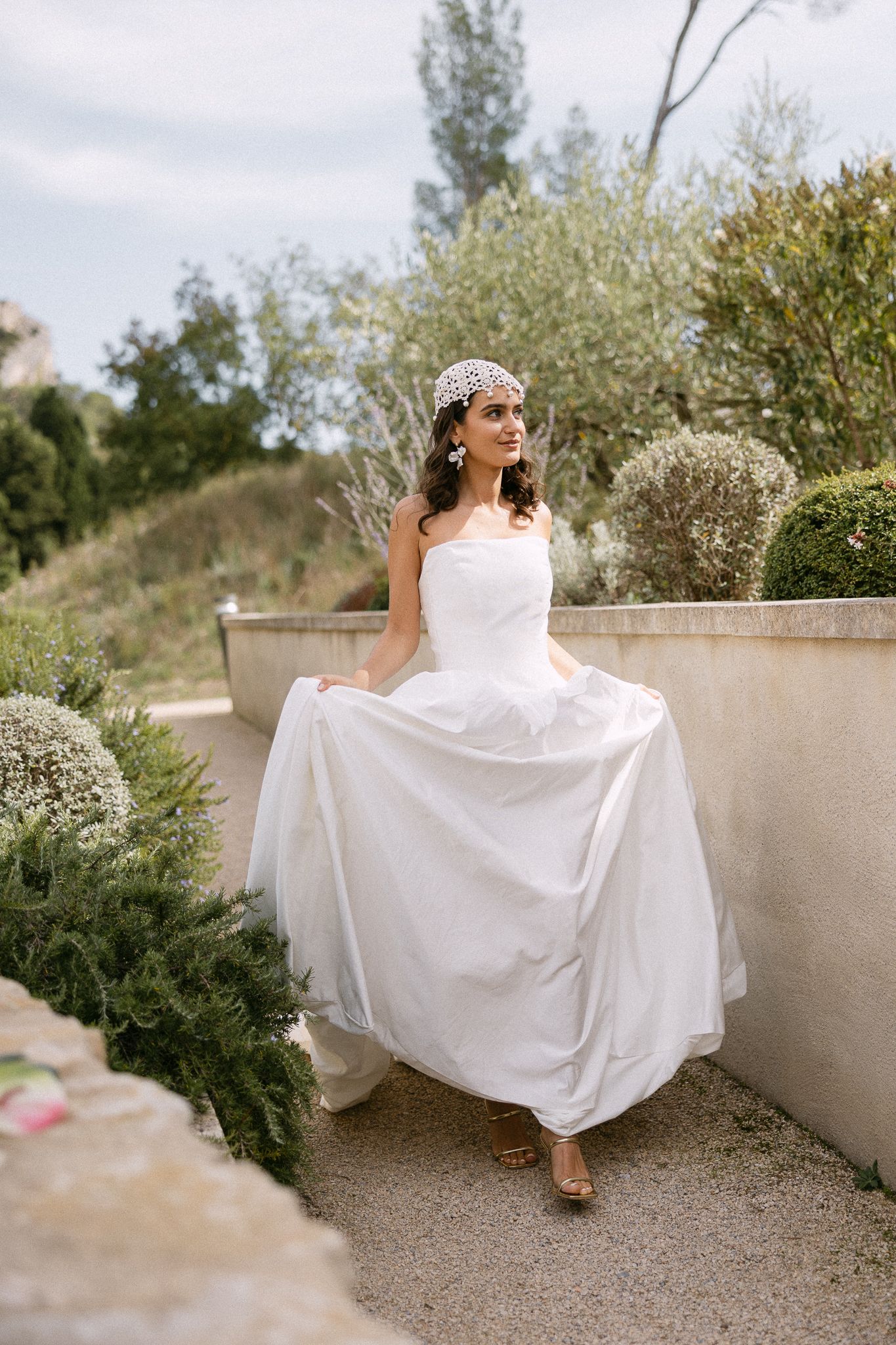Bride in white strapless gown and pearl headpiece twirling in a lush garden setting.