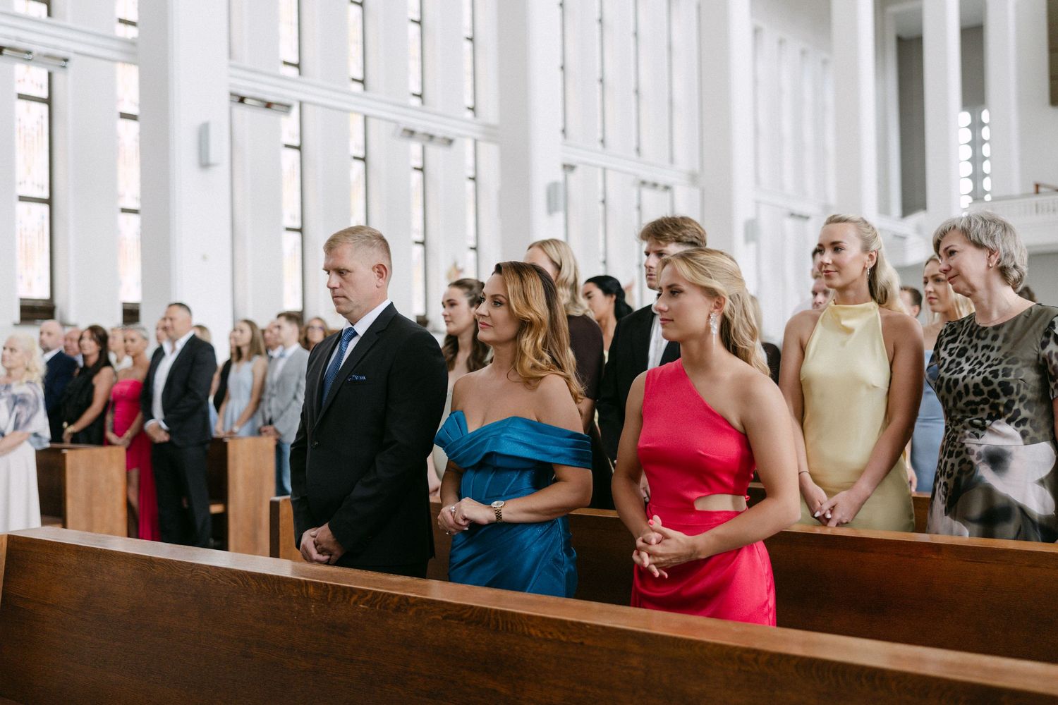 Wedding guests in colorful formal attire smile and stand during a church ceremony.
