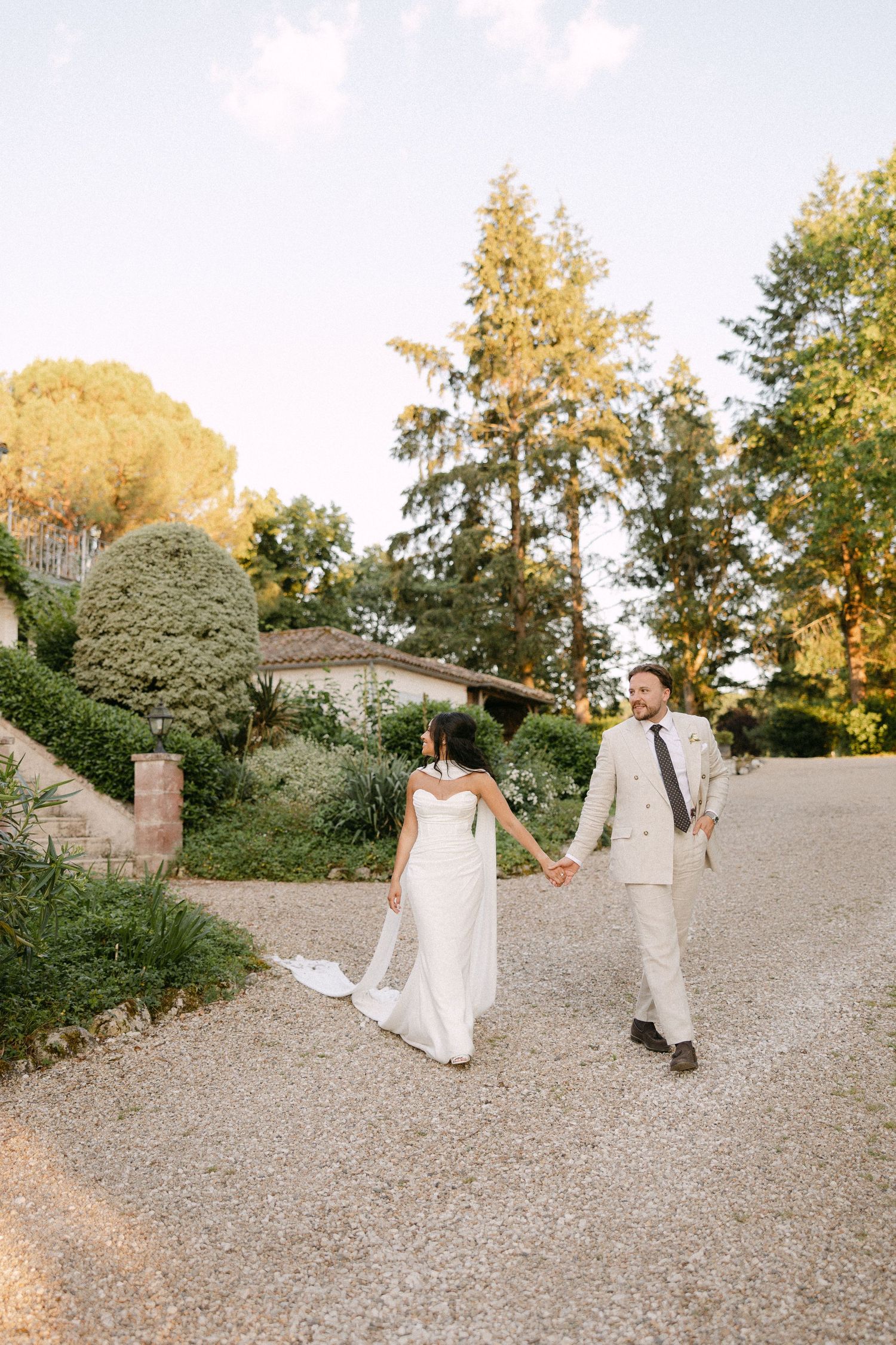 Bride and groom hold hands walking on a sunlit gravel path surrounded by lush trees and greenery.