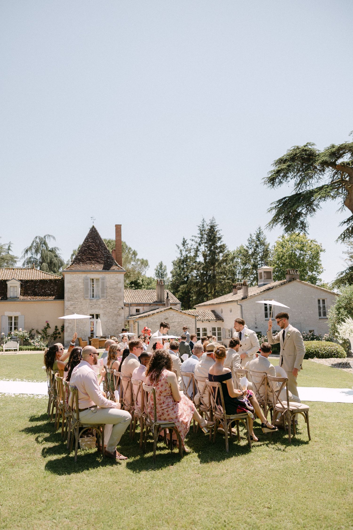 Outdoor wedding ceremony on green lawn with guests seated in wooden chairs before a charming French chateau on a sunny day.