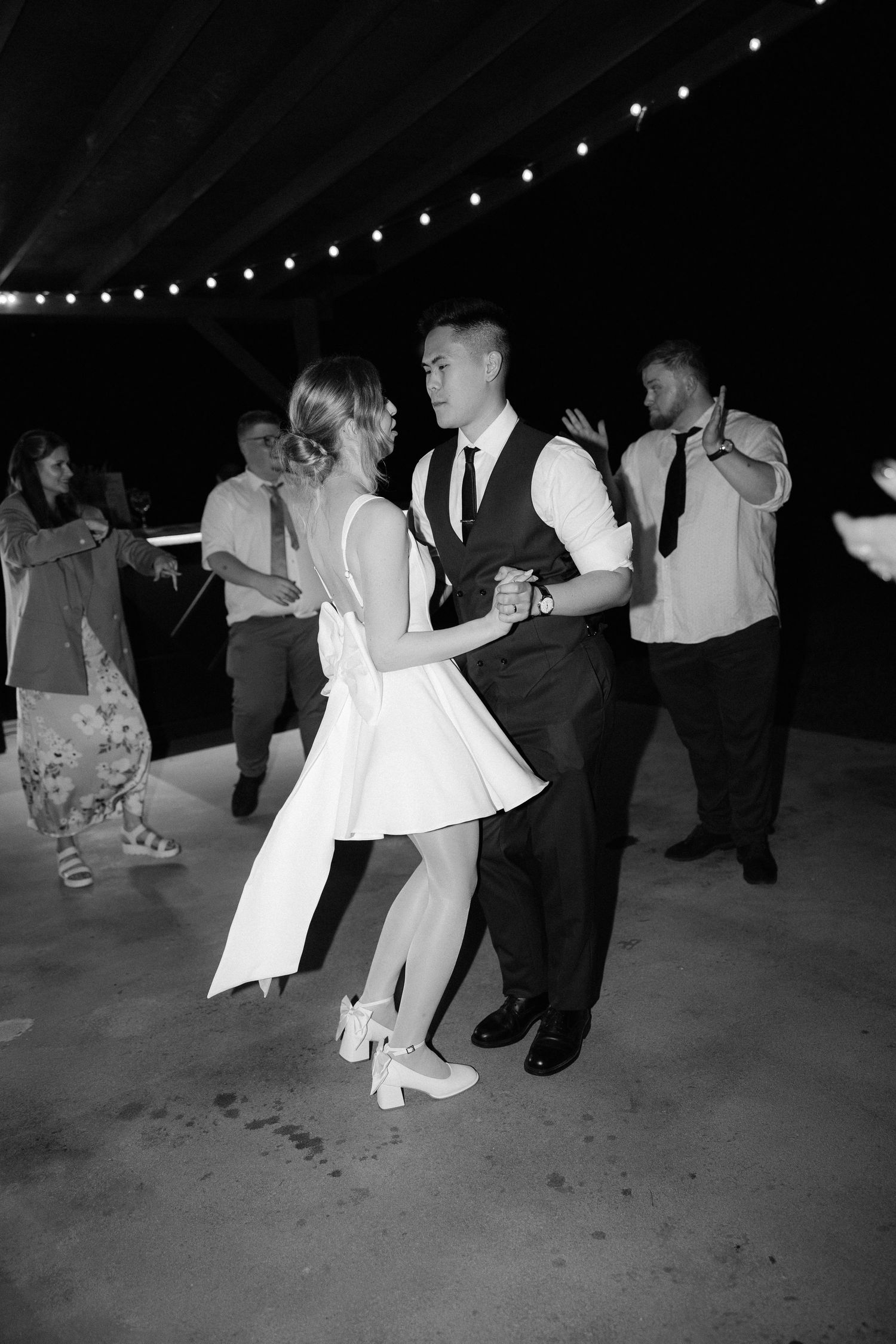 Bride and groom share first dance in black and white photo under string lights at night.