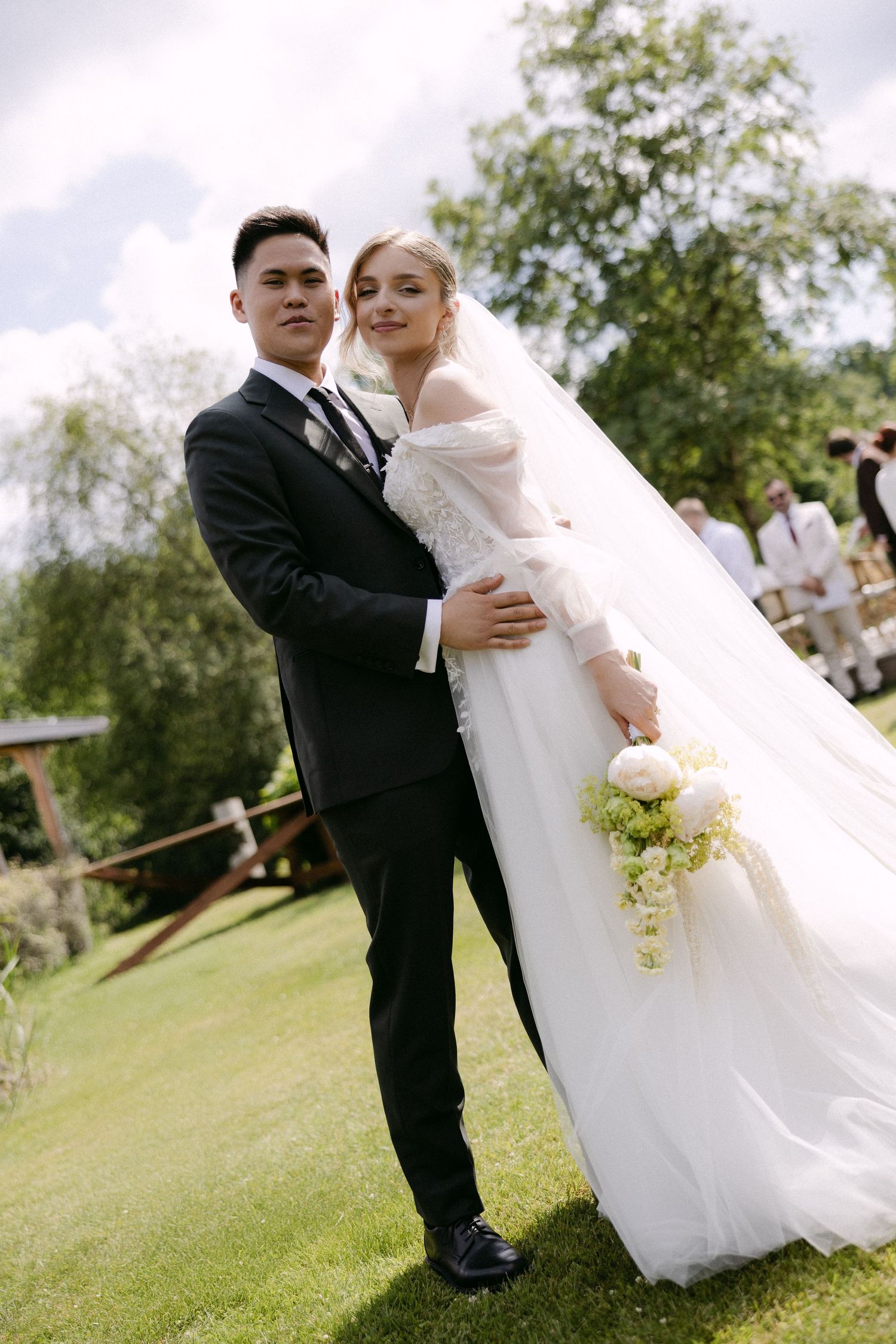 Bride and groom pose together on green lawn, she holds white peony bouquet, he wears black suit.