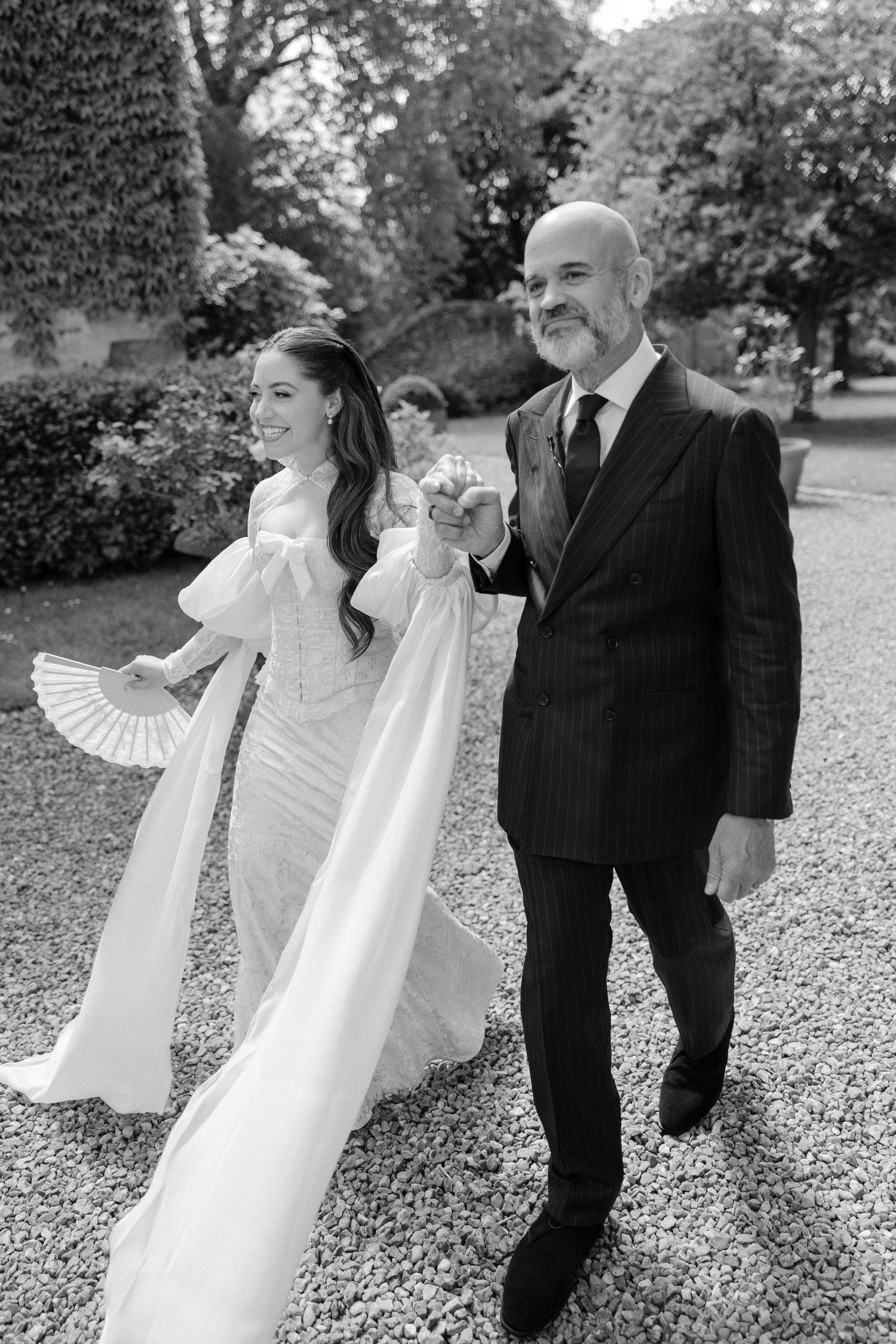 Black and white photo of smiling bride in lace gown walking with father of the bride outdoors.