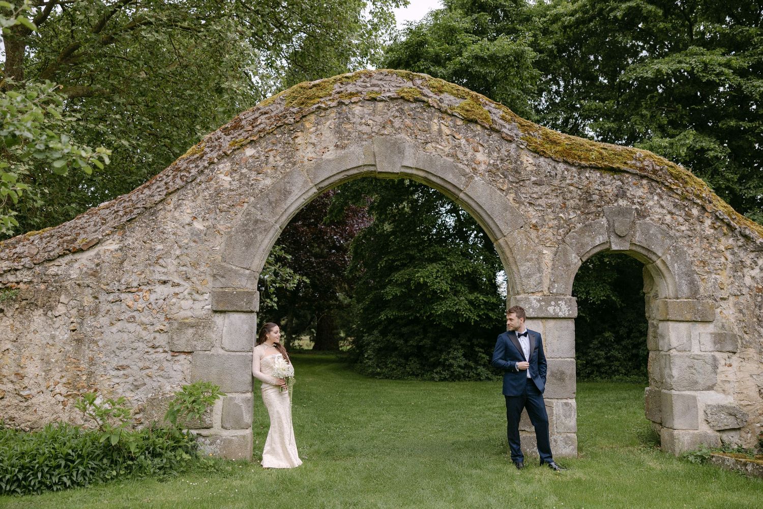 Bride and groom standing apart beneath a moss-covered stone arch in a lush green garden setting.