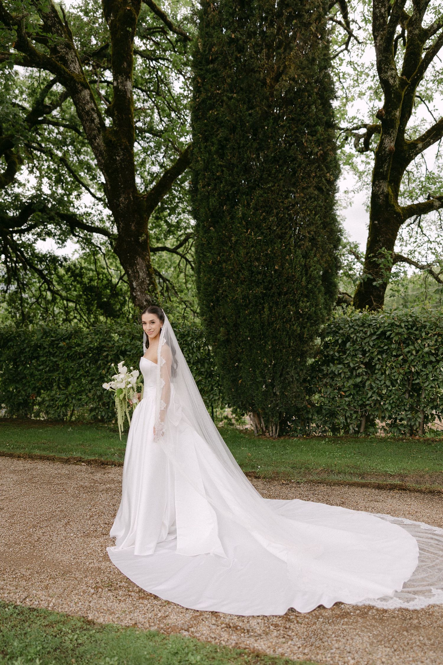 Bride in elegant white gown with long cathedral veil poses in lush garden with tall cypress trees.