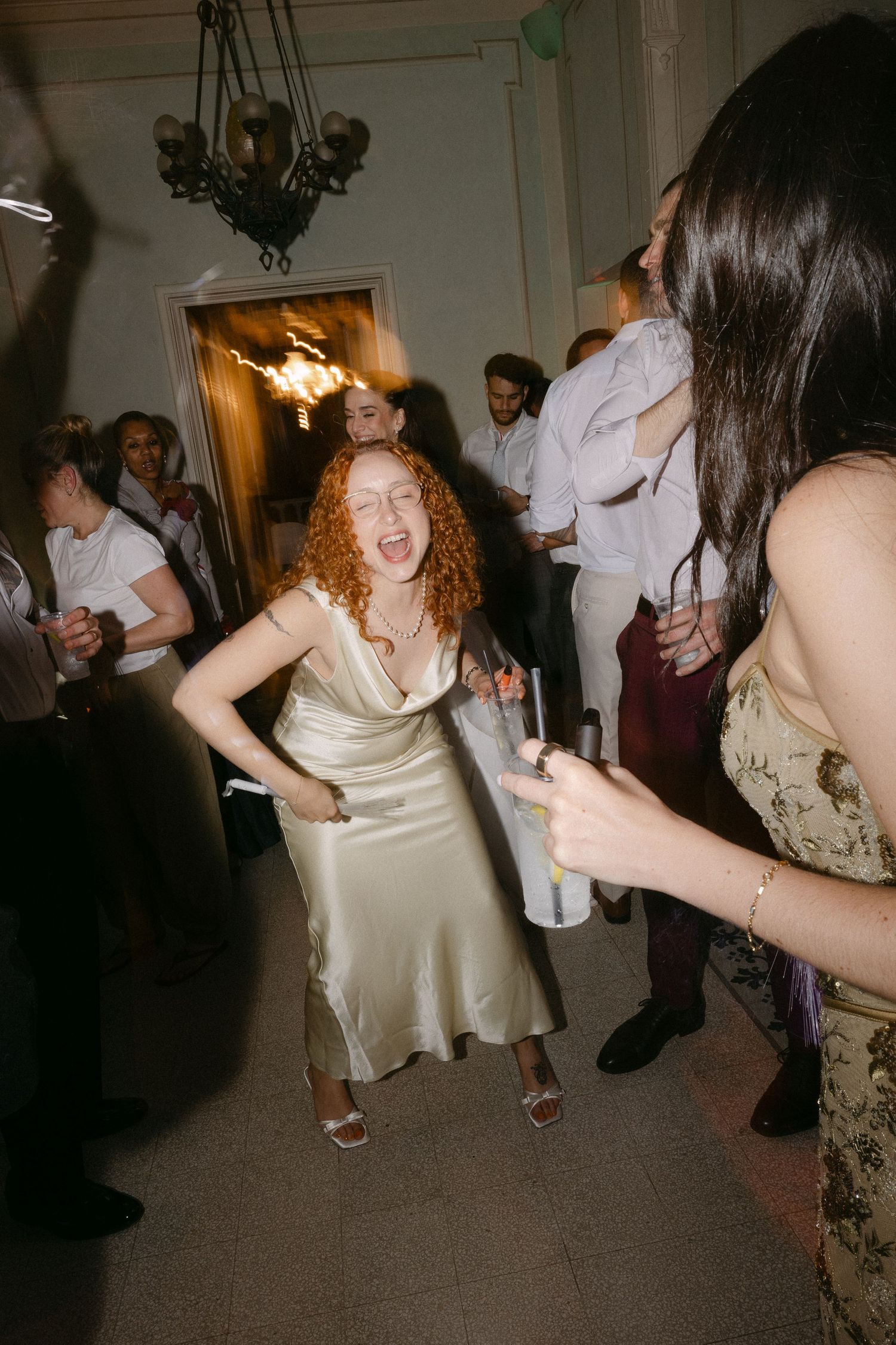 Bridesmaid in gold satin dress dancing energetically at a crowded wedding reception party.