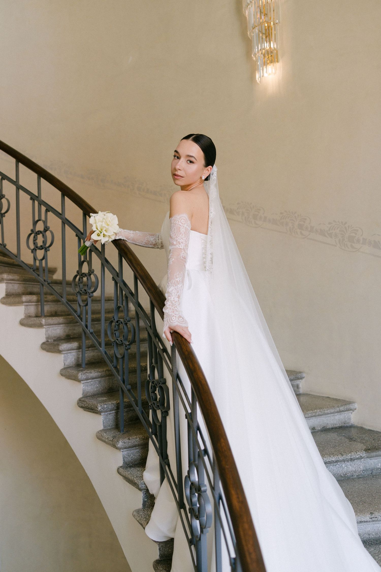 Beautiful bride in white ballgown with long veil descending ornate staircase holding floral bouquet.