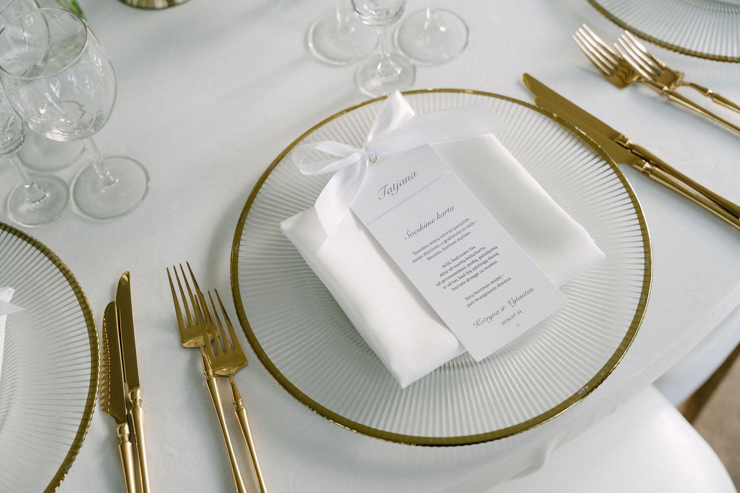 Elegant wedding table setting with gold cutlery, glass charger plate, and white menu card with ribbon.