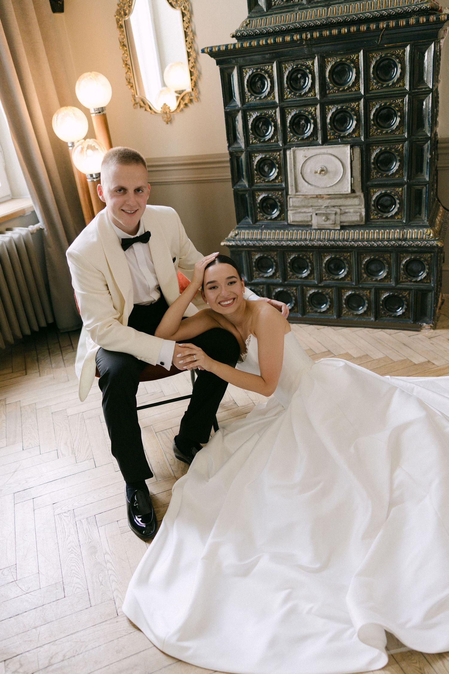 Smiling bride and groom in white tuxedo sit together by vintage fireplace on wedding day.