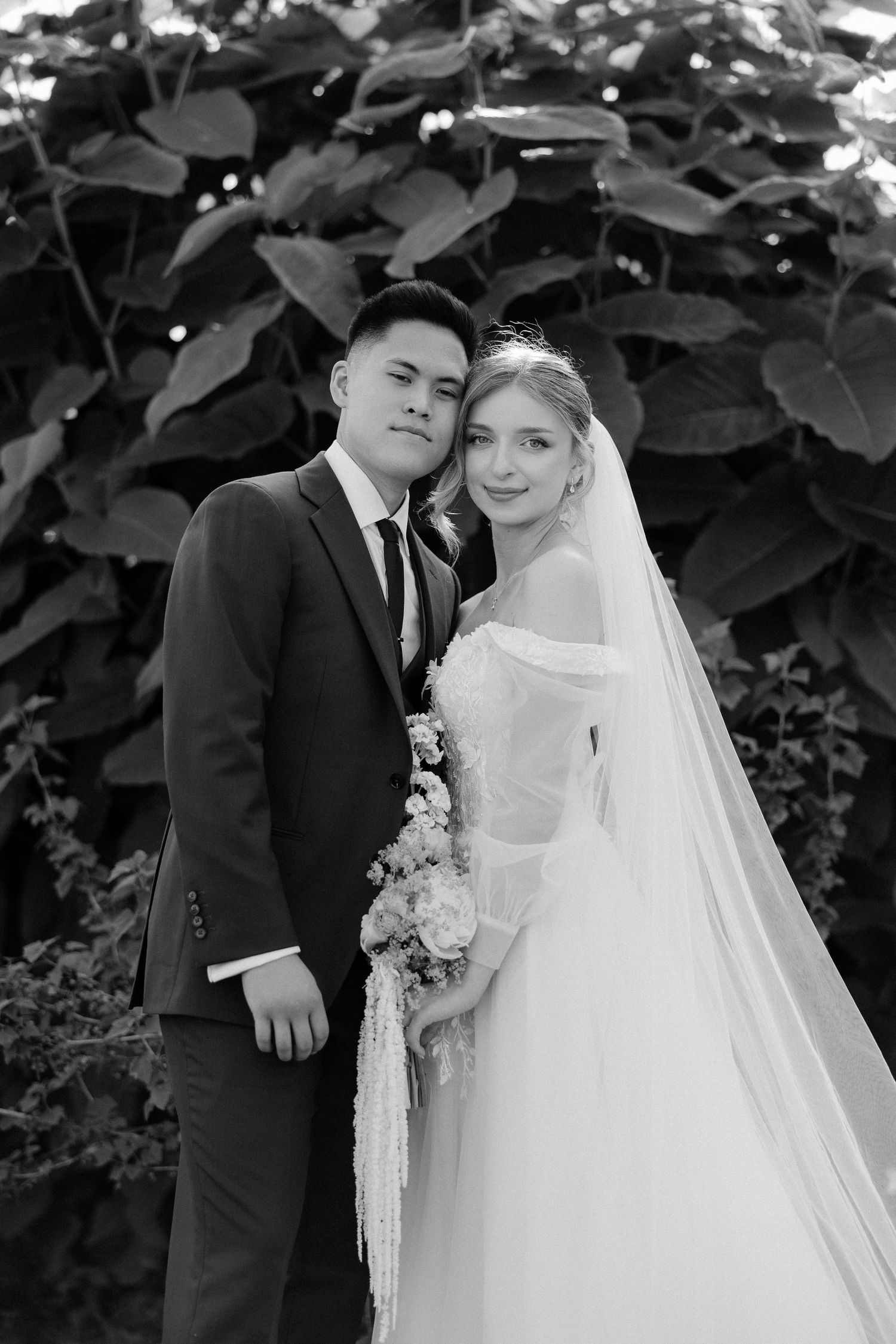 Black and white wedding portrait of bride and groom posing together in front of lush green foliage.