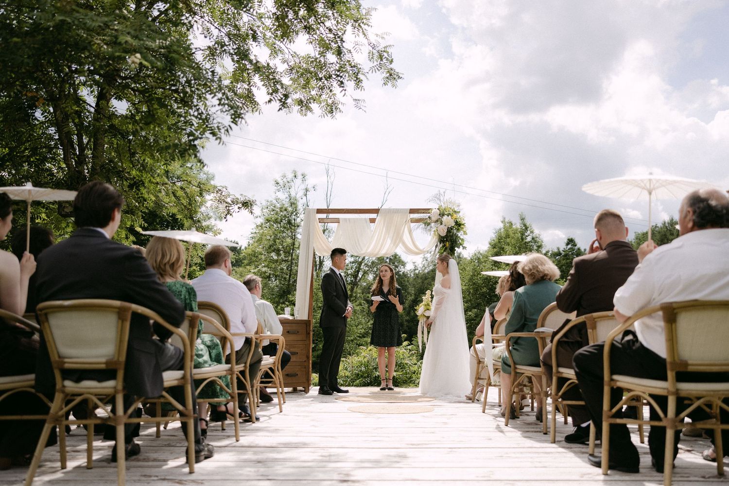 Outdoor wedding ceremony with guests seated in rows facing couple at floral arch altar.