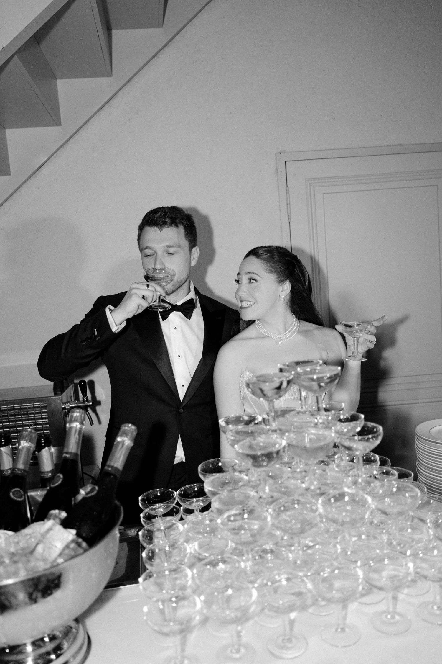 Newlyweds toast champagne beside a coupe glass tower at their black-tie wedding reception in black and white photo.