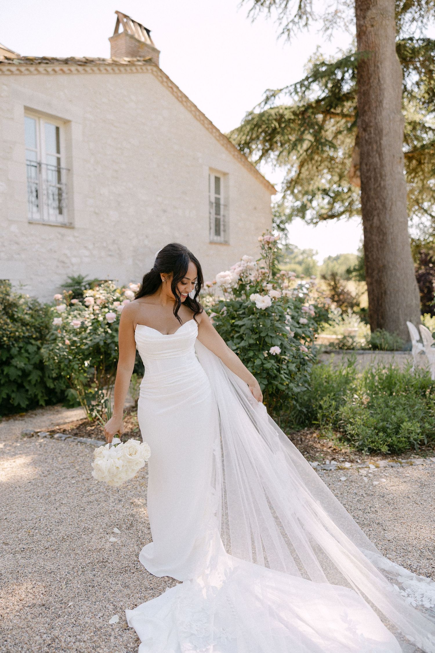 Radiant bride in strapless fitted gown with cathedral veil holding white bouquet outside French stone manor.