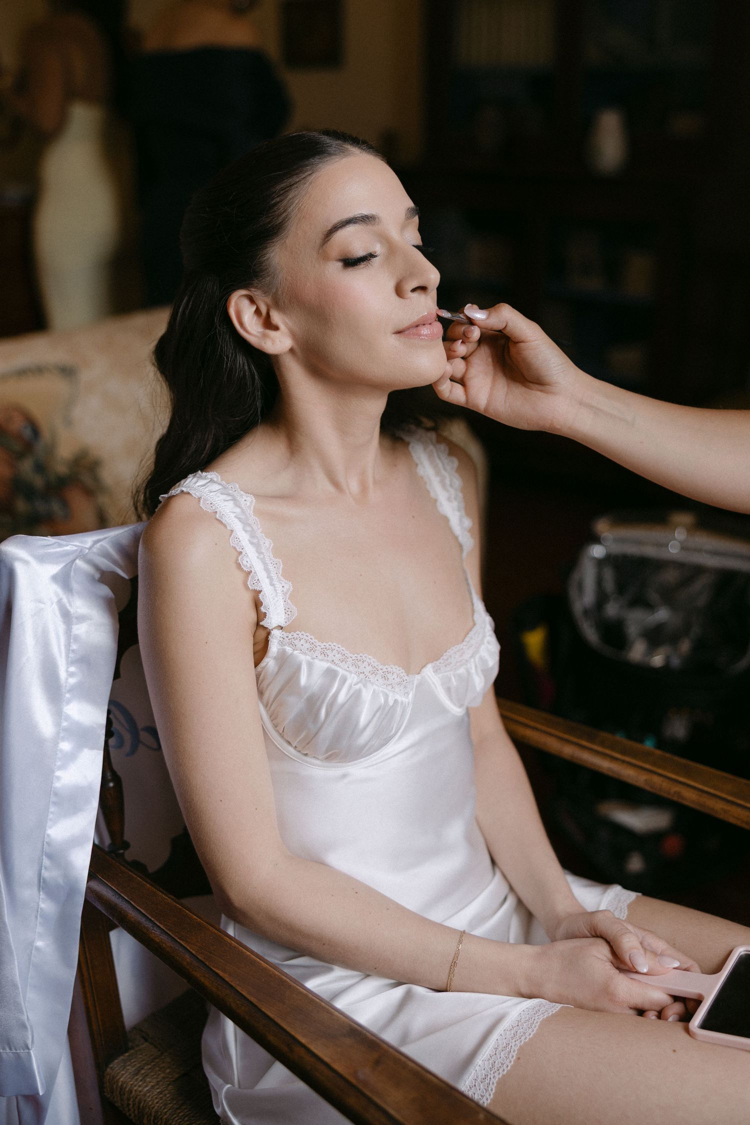 Bride in white satin slip dress gets makeup applied while seated in wooden chair before wedding.