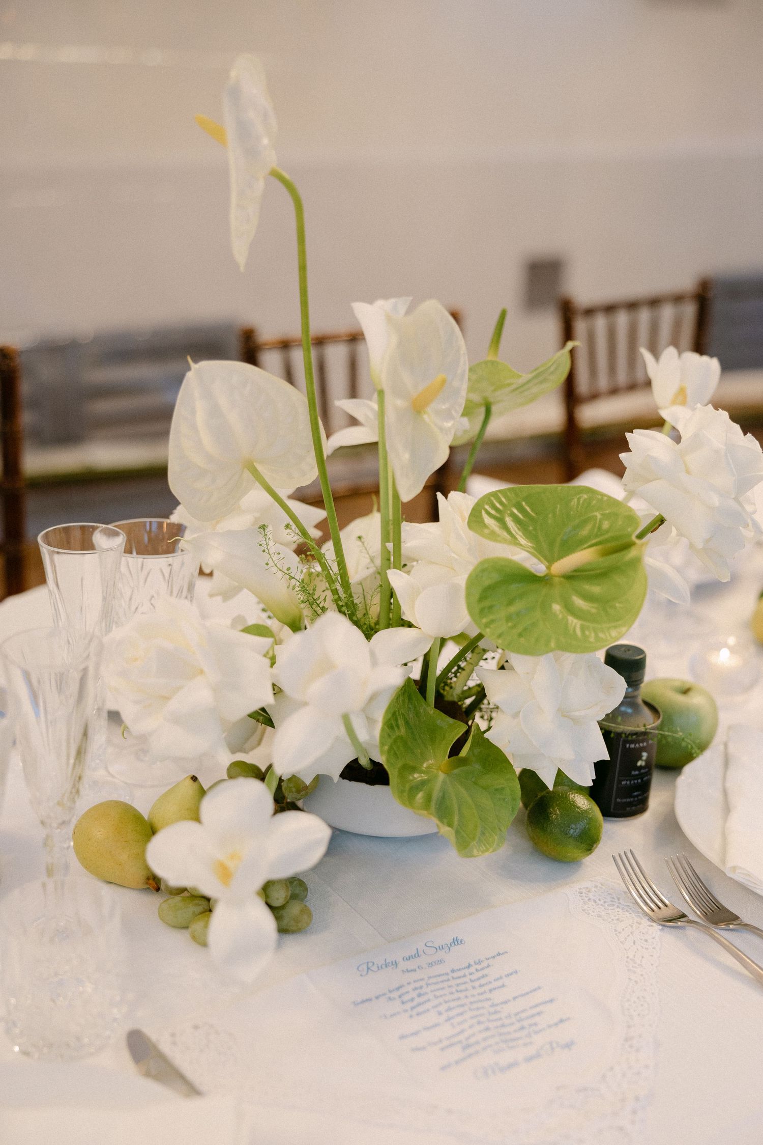 Elegant wedding table centerpiece with white anthuriums, green leaves, and fresh fruit on white linen.