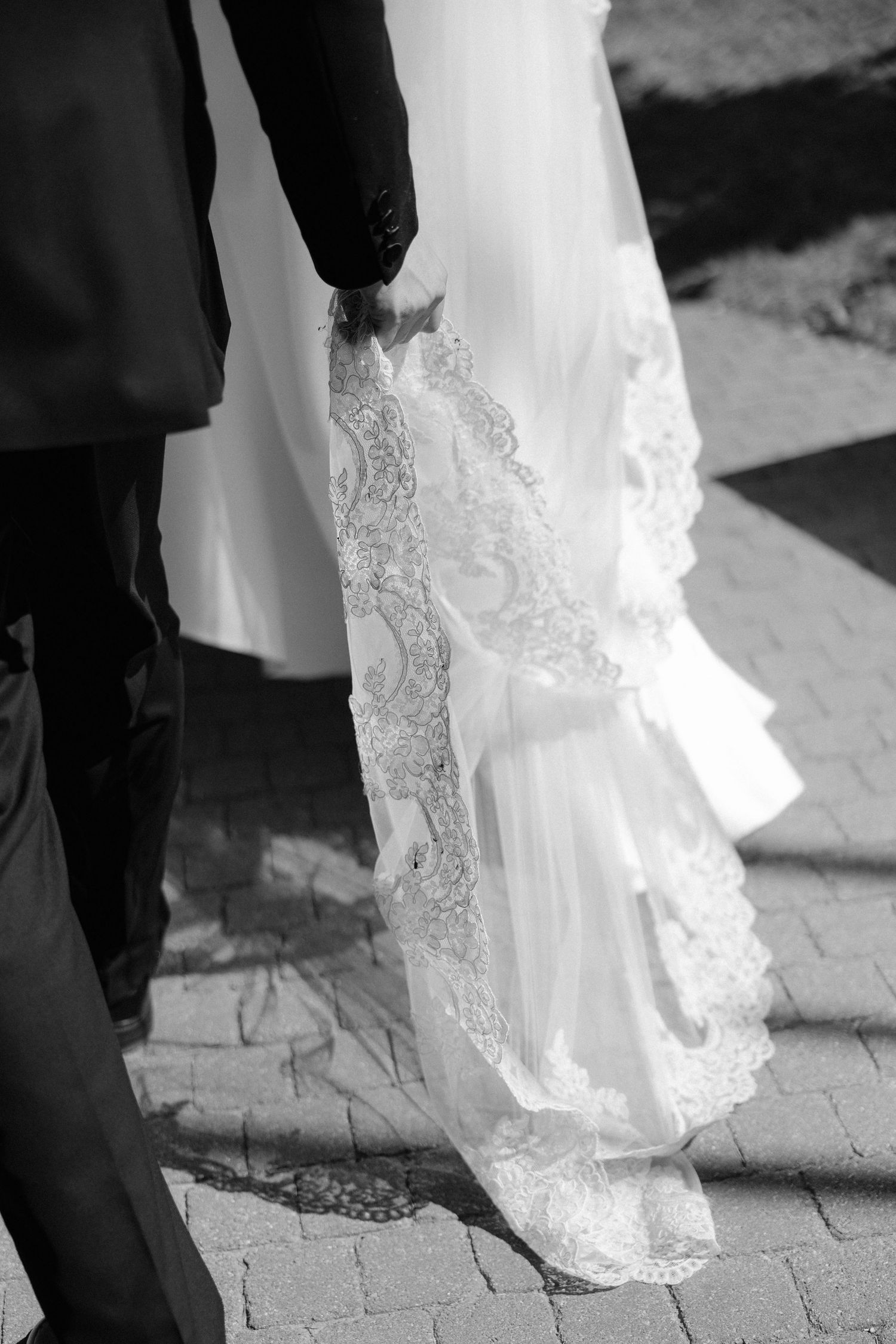 Black and white photo of a groom holding the lace-trimmed veil of a bride walking on cobblestones.