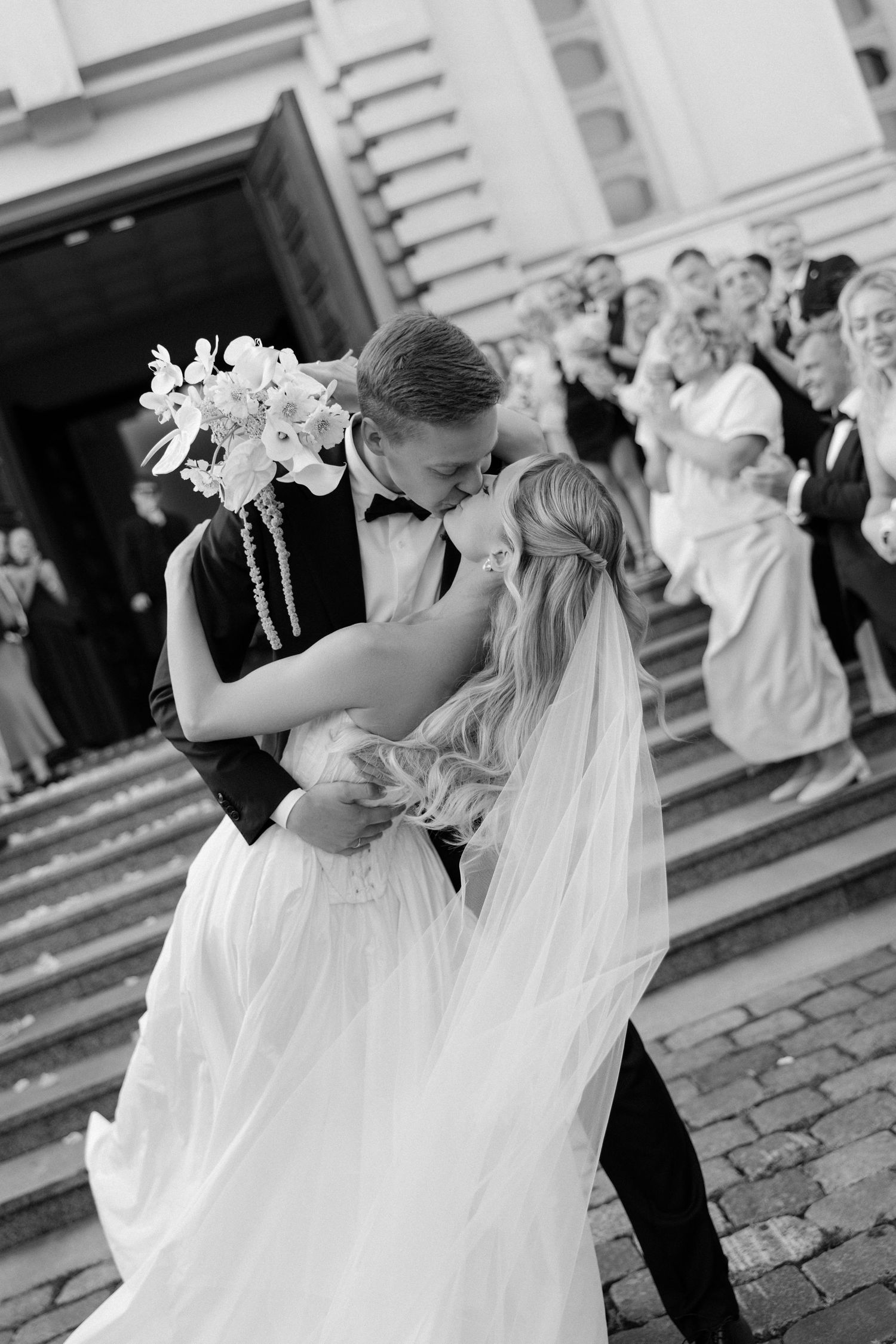 Black and white photo of bride and groom sharing a kiss outside a church with wedding guests on steps behind them.
