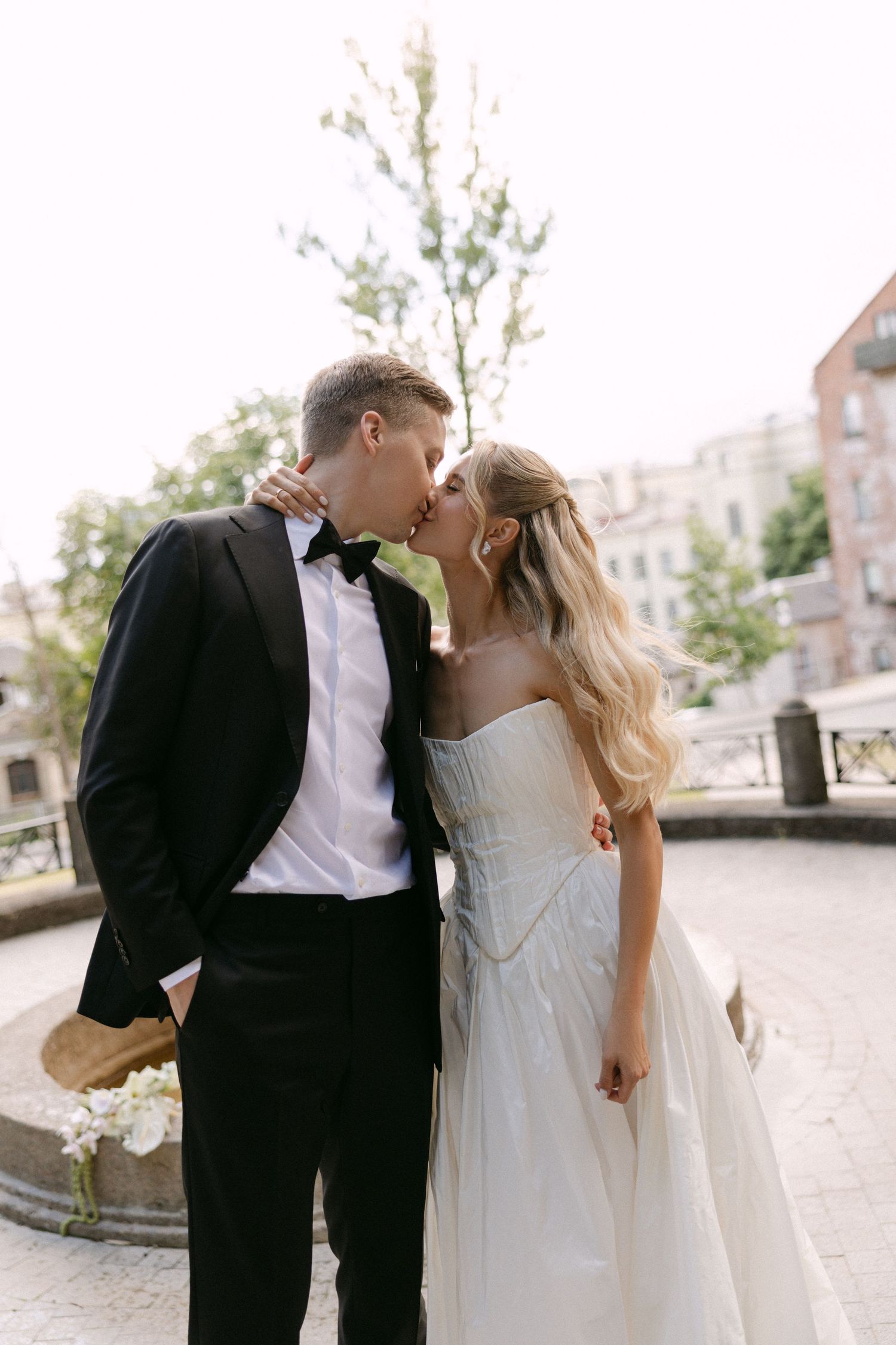 Elegant bride and groom share a tender kiss outdoors near a fountain in classic black tie wedding attire.