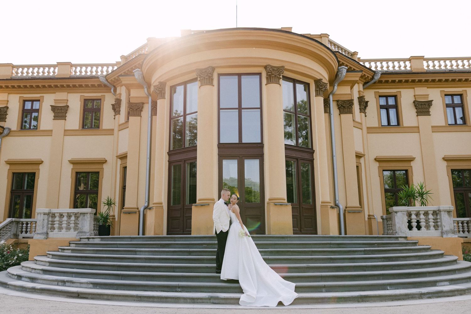 Elegant bride and groom embracing on grand steps of a classical mansion wedding venue in golden sunlight.