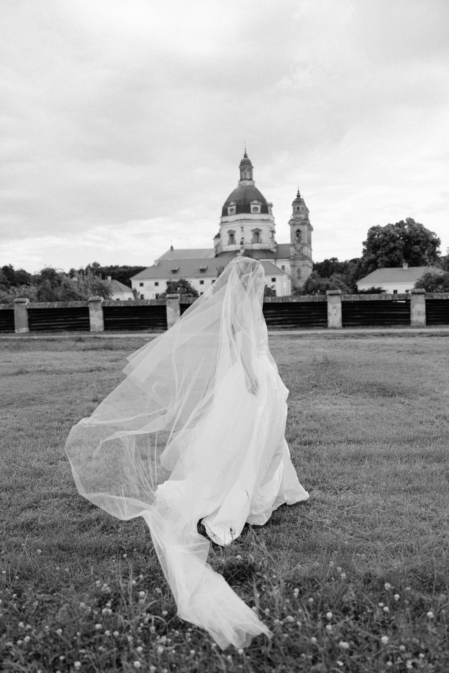 Black and white photo of bride in flowing gown and veil standing before a historic baroque church.