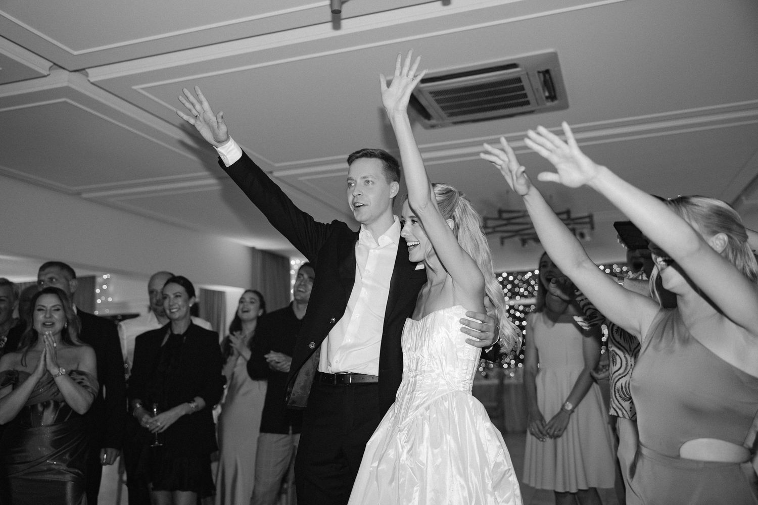 Bride and groom celebrate joyfully with raised hands on the dance floor surrounded by wedding guests.
