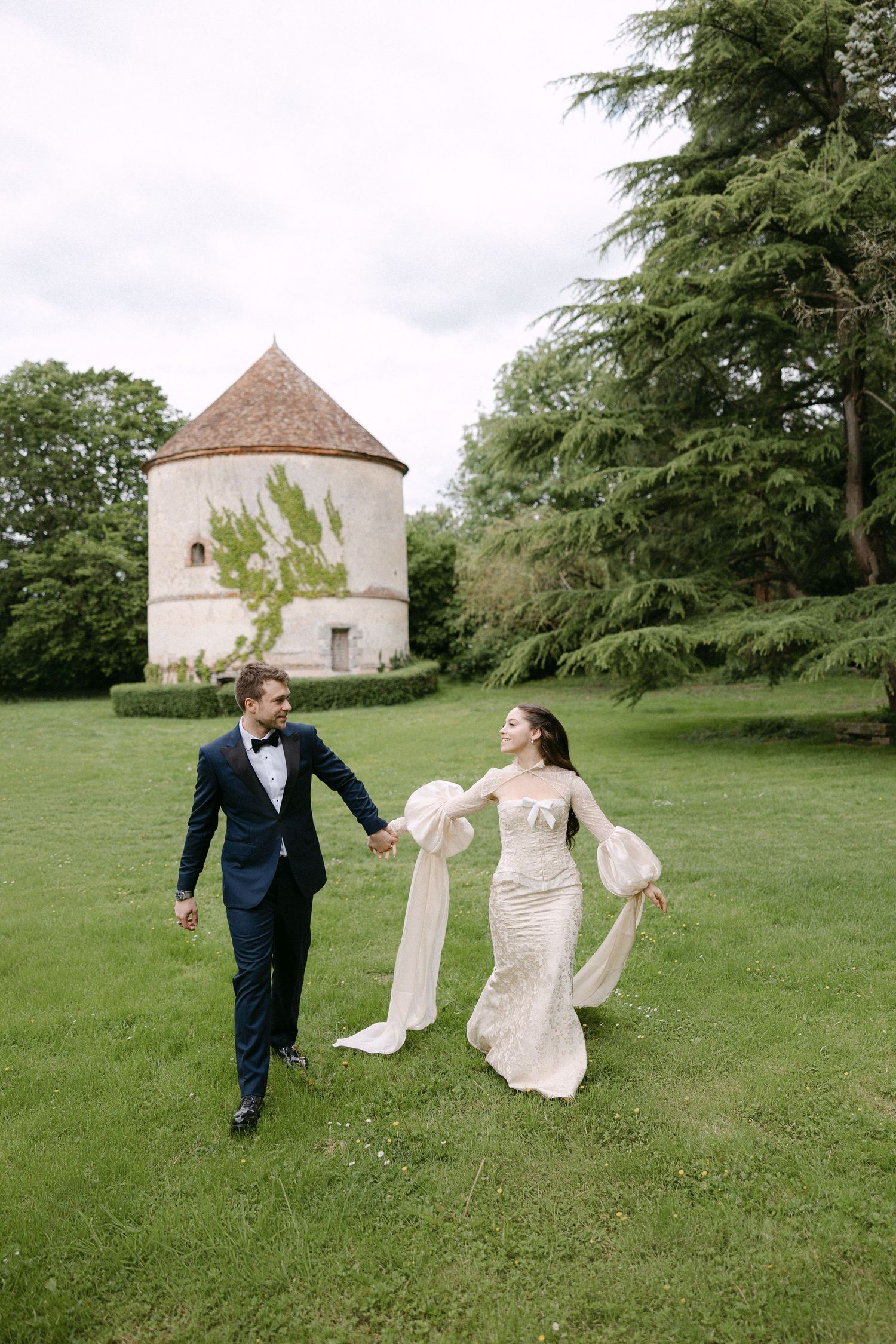 Bride and groom hold hands walking on green lawn with historic round stone tower and trees in background.