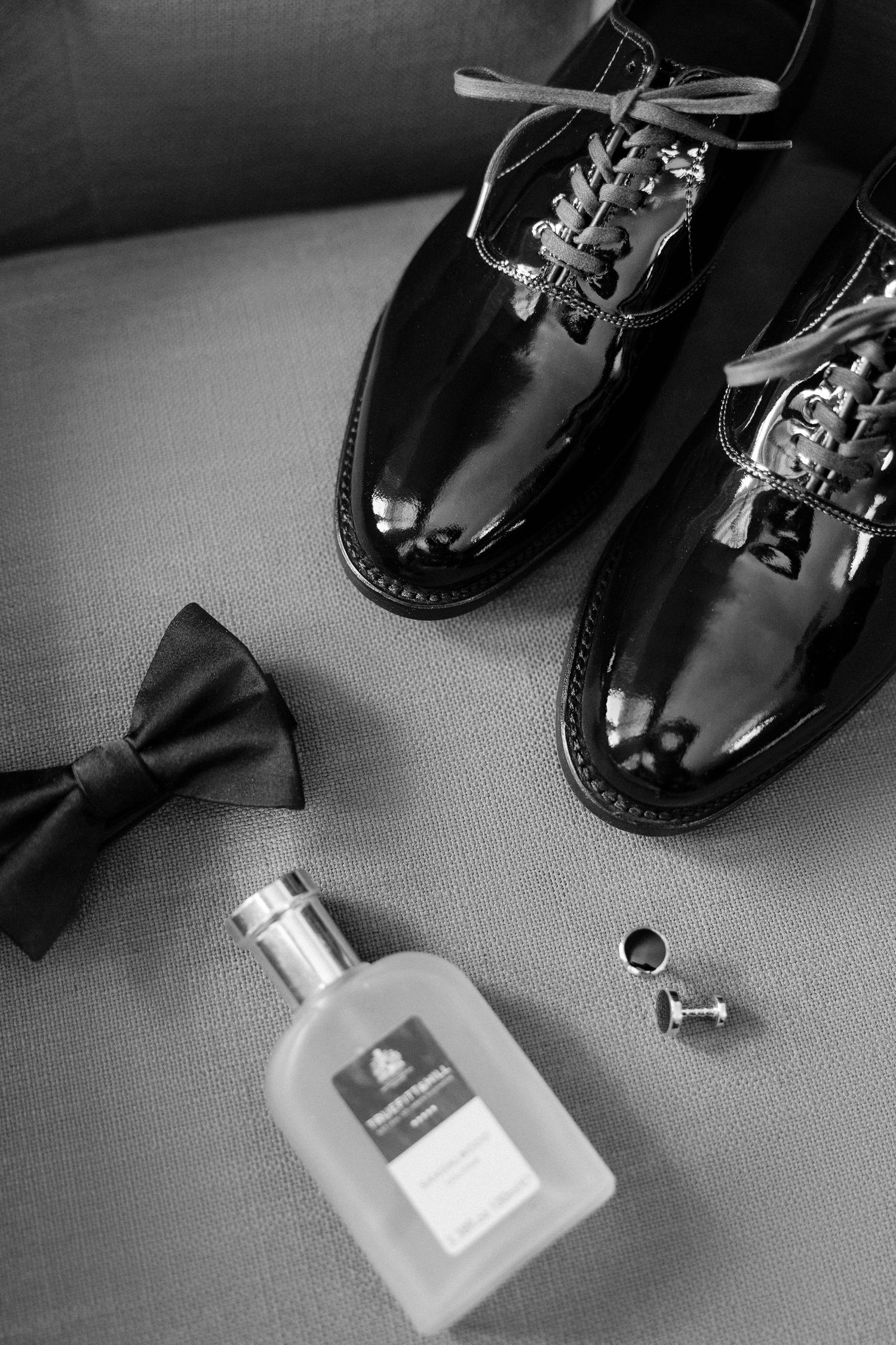Black and white flat lay of grooms patent leather oxford shoes, bow tie, cologne, and cufflinks.