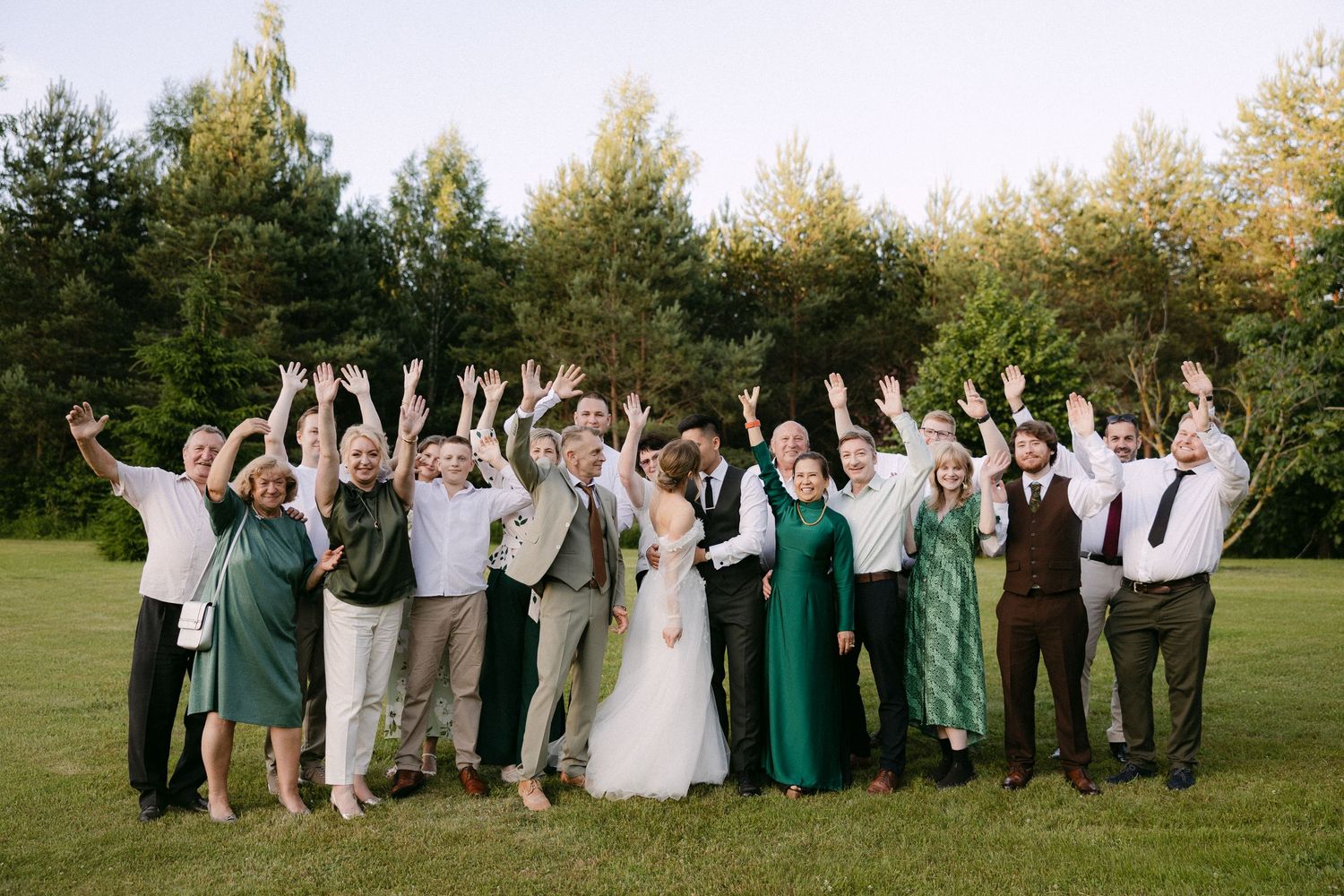 Joyful wedding party group photo with raised hands on green lawn surrounded by tall pine trees.