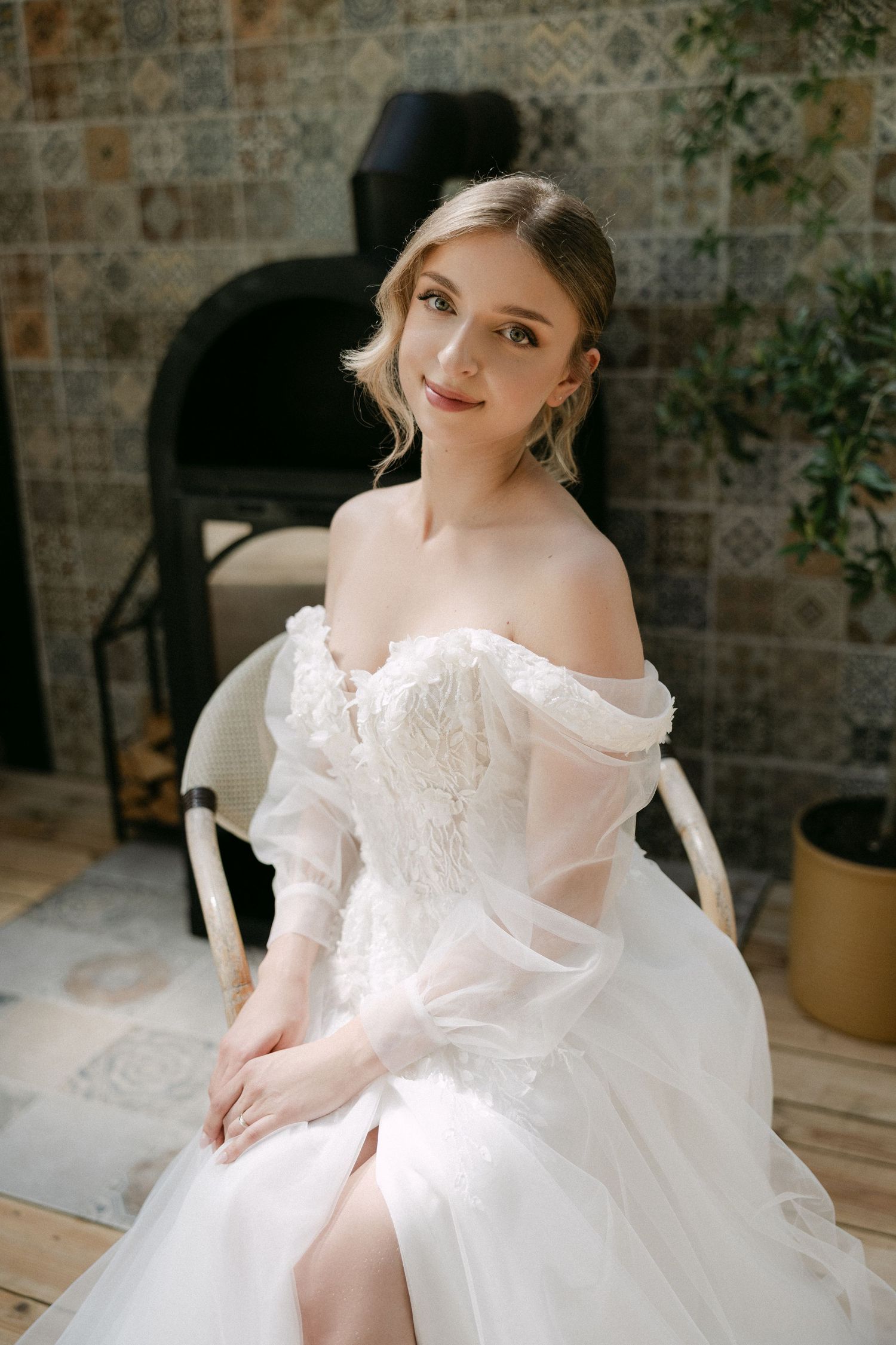 Beautiful bride in off-shoulder white tulle wedding dress sitting and smiling near tiled fireplace.