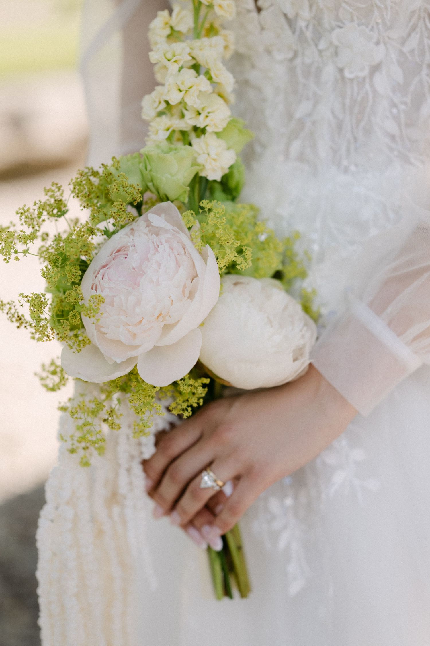 Bride holds elegant white peony and green floral bouquet, diamond ring visible on her finger.