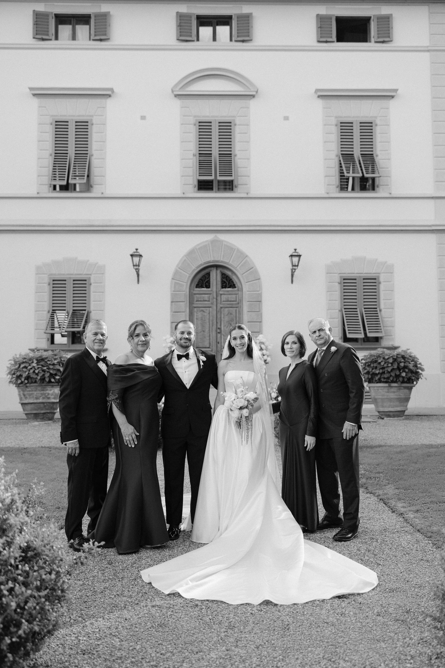 Black and white portrait of bride, groom and family posing before a classic Italian villa facade.