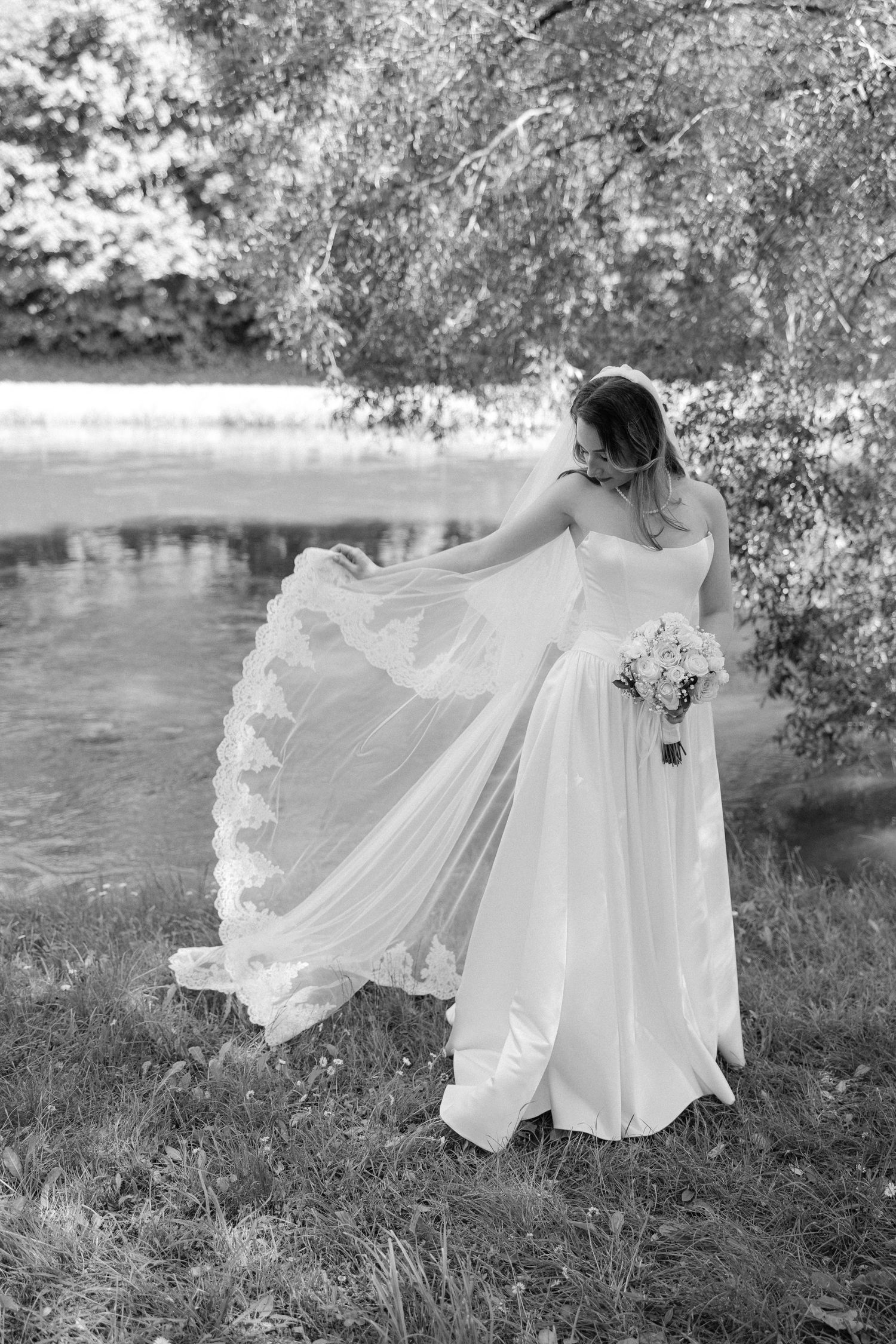 Black and white photo of bride in flowing wedding dress twirling near a peaceful lakeside setting.