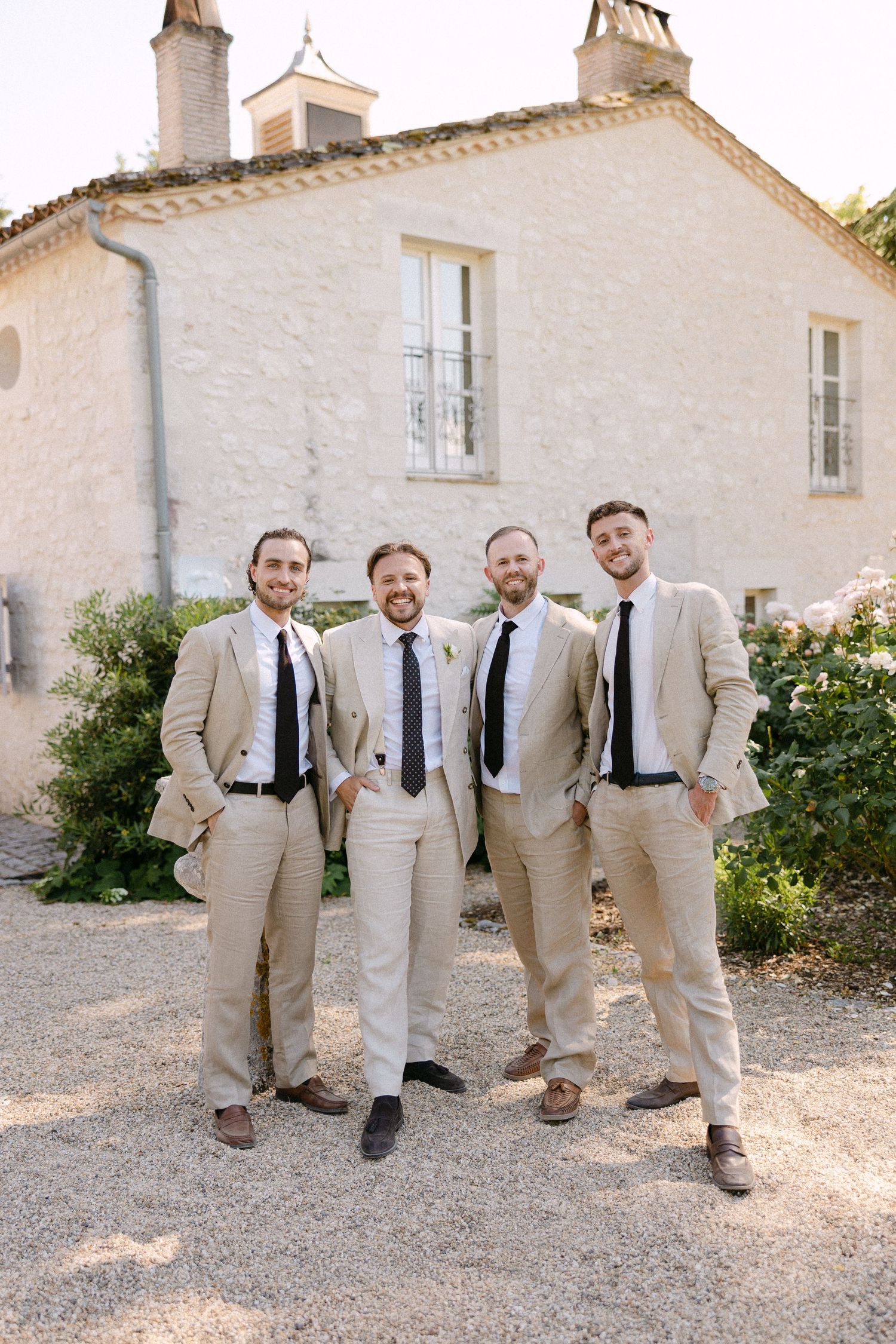 Four groomsmen in matching beige suits and dark ties pose together outside a rustic stone building.
