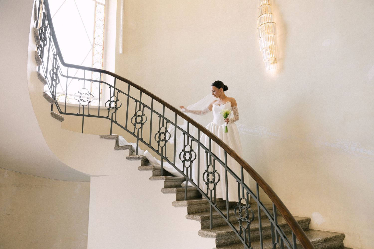 Bride in white gown descending elegant curved staircase with ornate iron railing in a bright, classic venue.