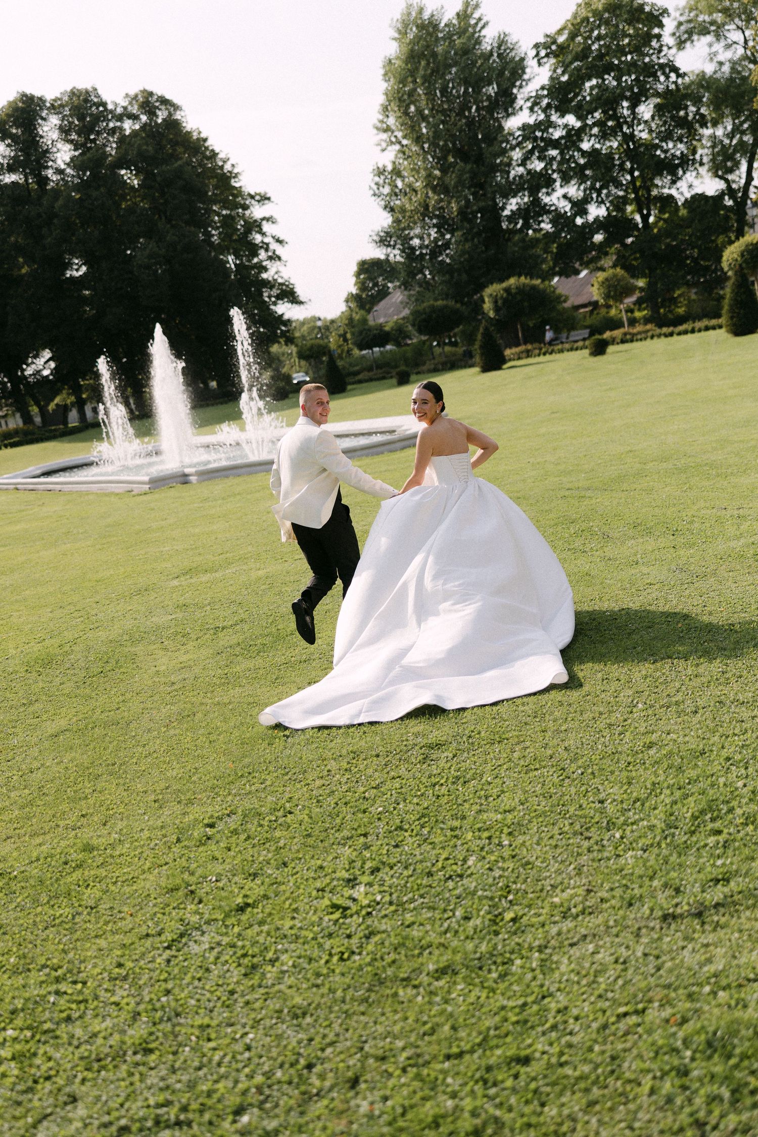 Bride and groom running hand in hand across lush green lawn with fountain backdrop on their wedding day.