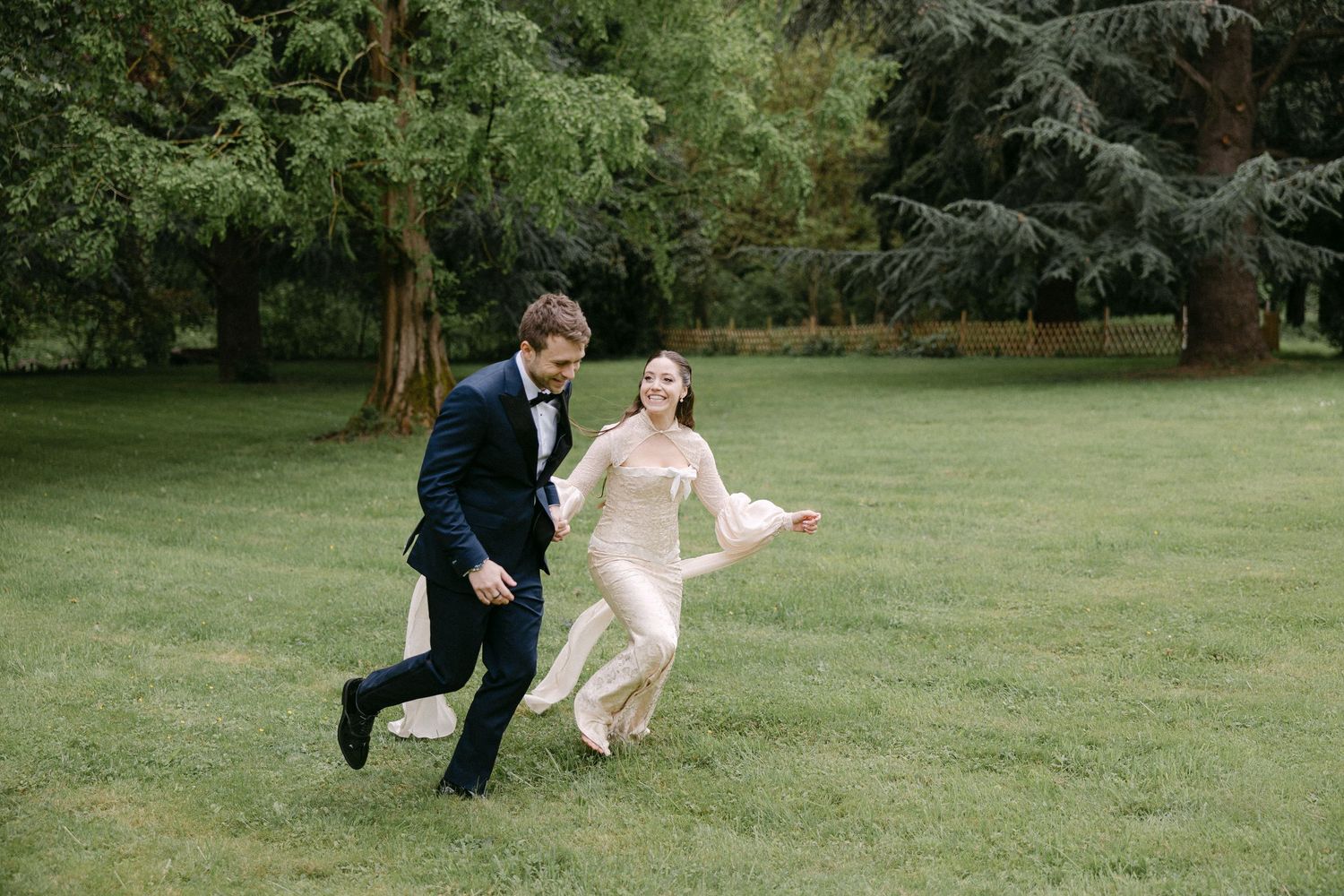 Joyful bride and groom running playfully across green lawn with tall evergreen trees in background.