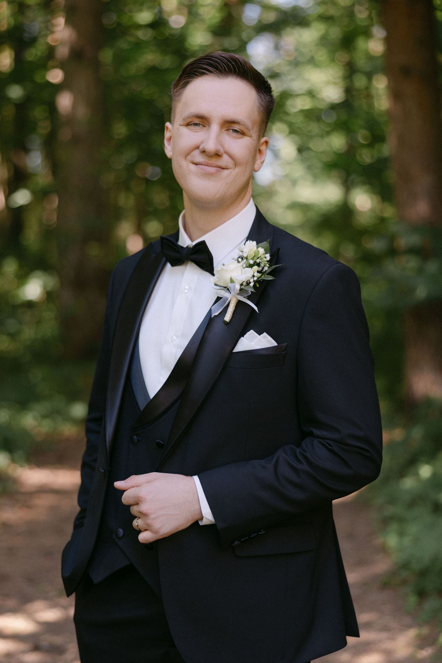 Dapper groom in black tuxedo with bow tie and floral boutonniere smiling in a sunlit forest.