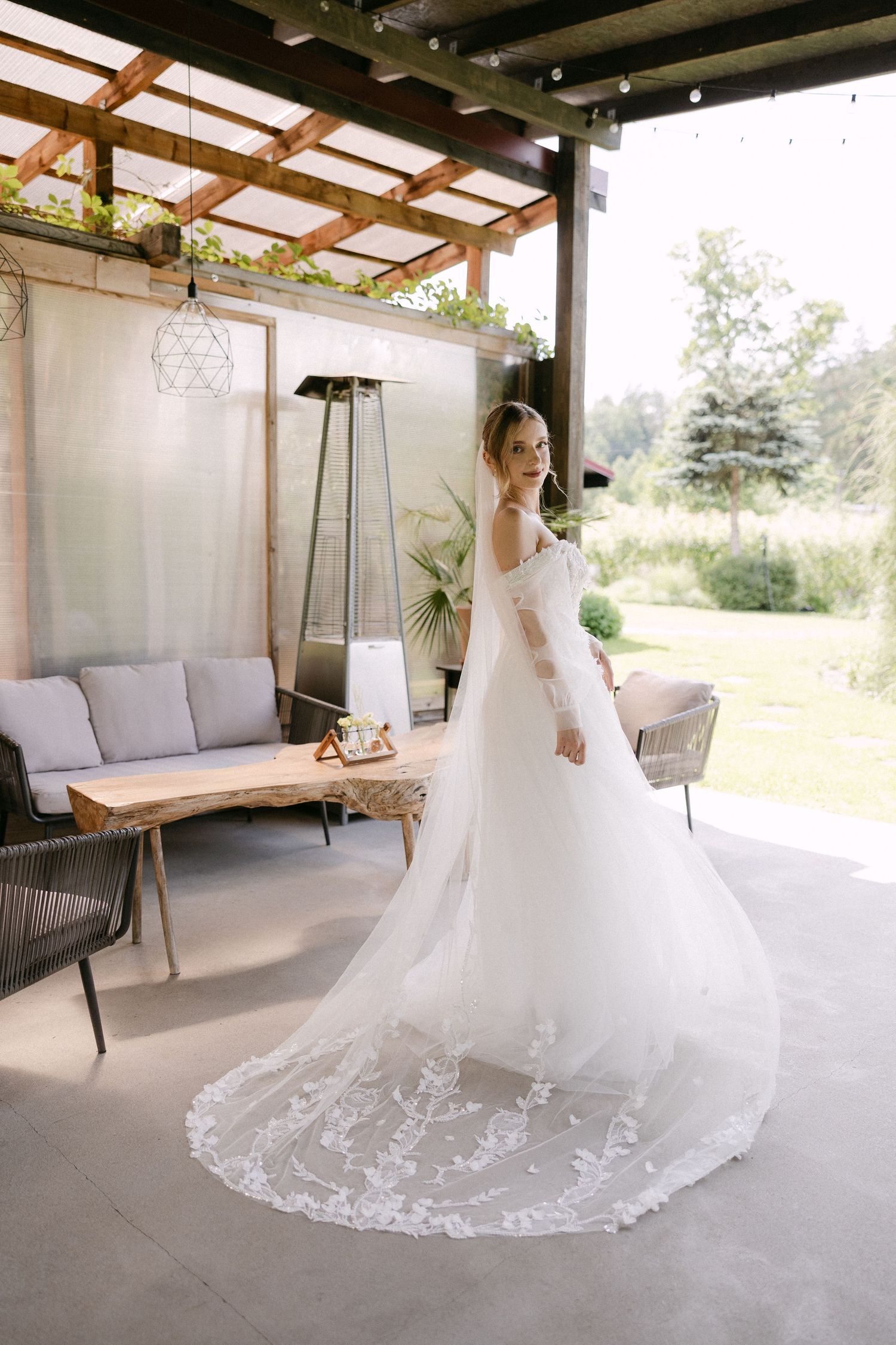 Bride in elegant lace wedding gown with long cathedral veil posing on outdoor patio with garden view.